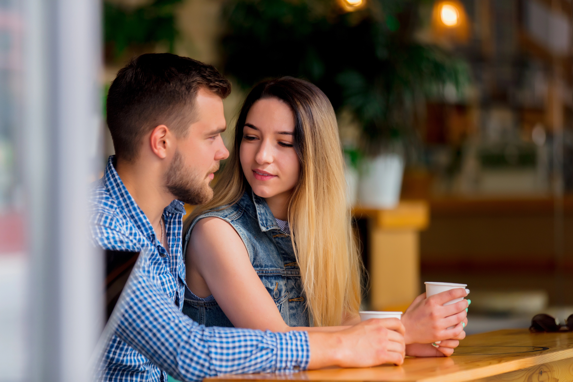 A man and woman sit closely together at a wooden table in a cozy café. The man, with short hair and a beard, is wearing a blue checkered shirt. The woman, with long, straight hair, wears a sleeveless denim jacket. Both are holding white coffee cups, smiling softly.