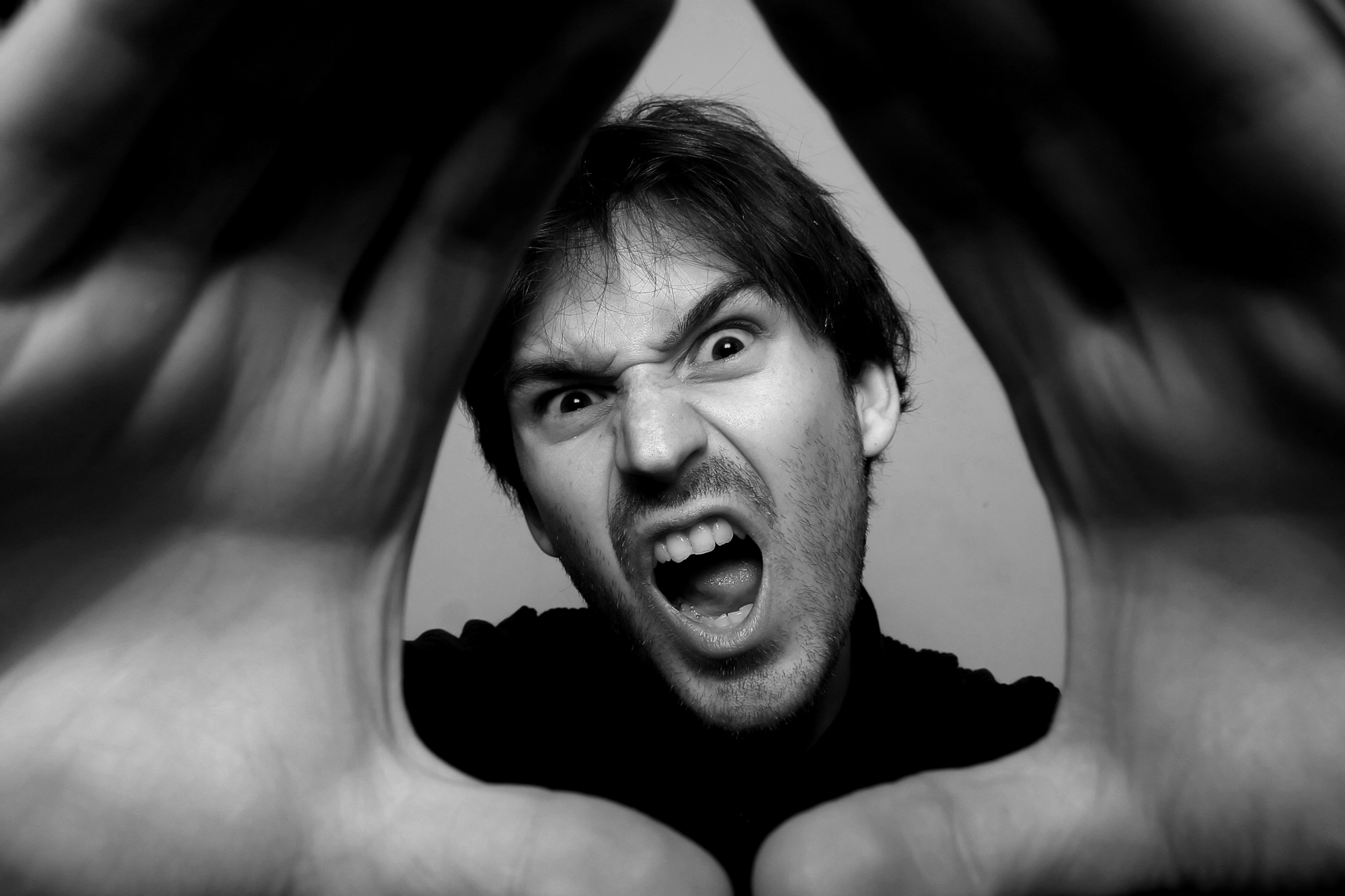 Black and white close-up of a person with an intense expression, mouth open and teeth showing, appearing to shout. Their hands are in front, framing their face in a triangular shape. The emotions conveyed are strong and dramatic.