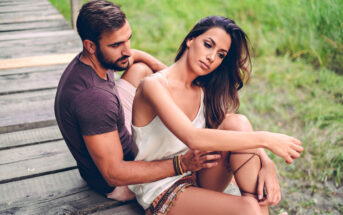 A man and a woman sit on a wooden pathway in an outdoor setting with greenery around. The man, wearing a dark T-shirt, is seated behind the woman, who is in a white top and looking contemplative, with her arm resting on her knee.