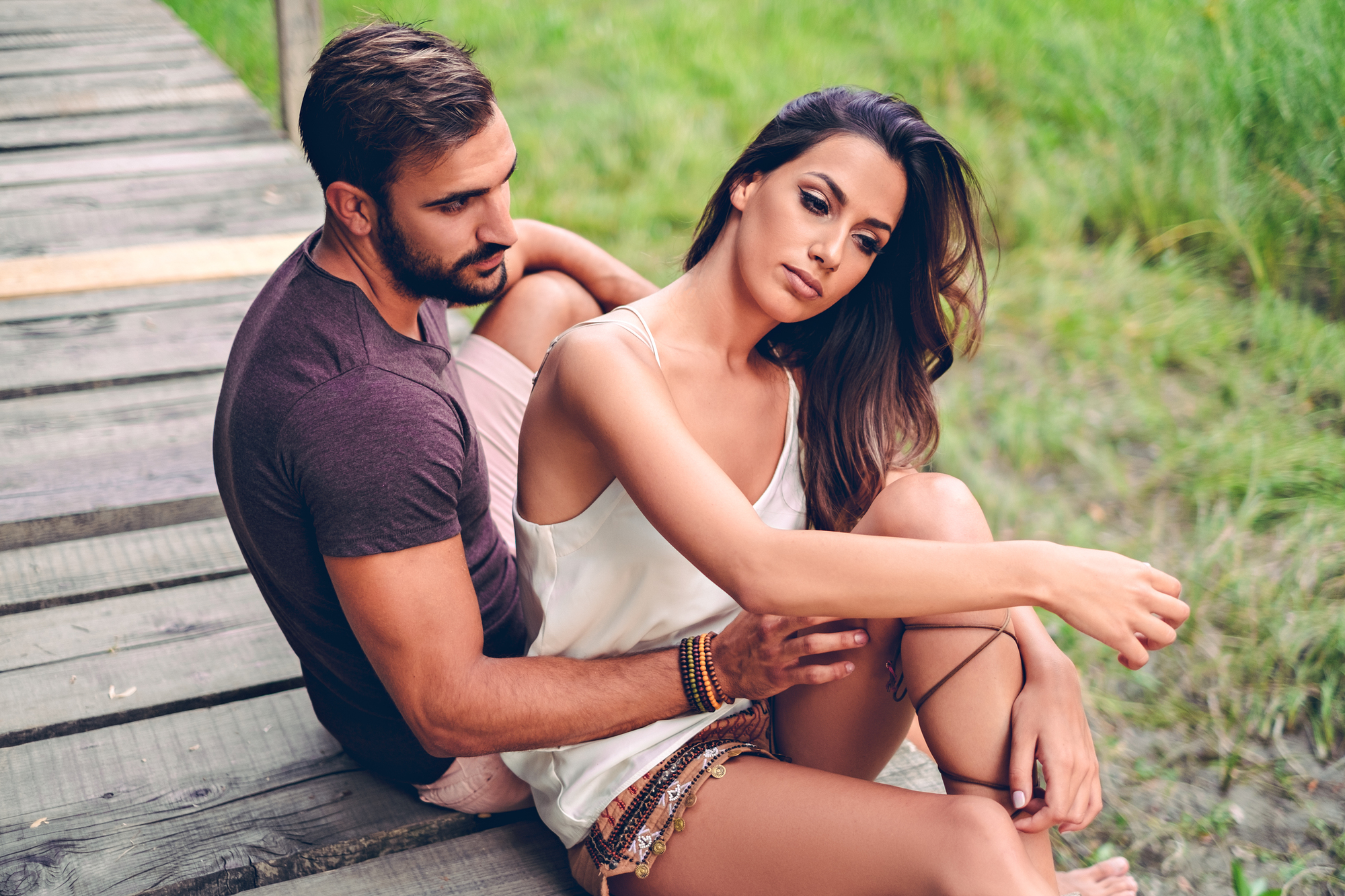 A man and a woman sit on a wooden pathway in an outdoor setting with greenery around. The man, wearing a dark T-shirt, is seated behind the woman, who is in a white top and looking contemplative, with her arm resting on her knee.