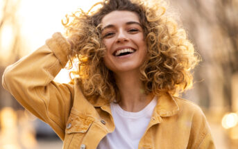 A young woman with a joyful expression stands outdoors, smiling brightly. She has curly hair and is wearing a yellow denim jacket over a white shirt. Her hand is playfully touching her hair, and the background is softly blurred with warm sunlight.