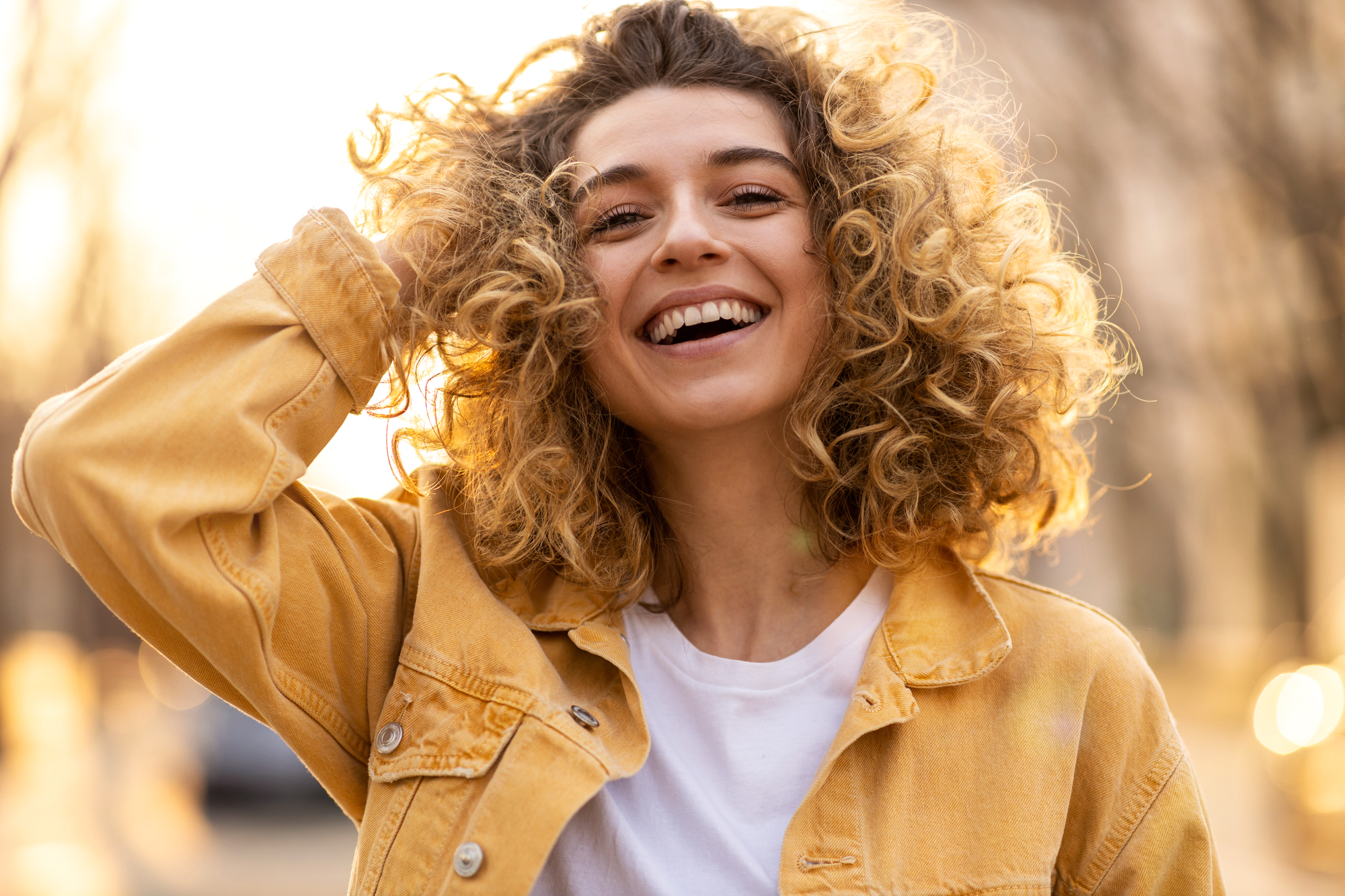 A young woman with a joyful expression stands outdoors, smiling brightly. She has curly hair and is wearing a yellow denim jacket over a white shirt. Her hand is playfully touching her hair, and the background is softly blurred with warm sunlight.