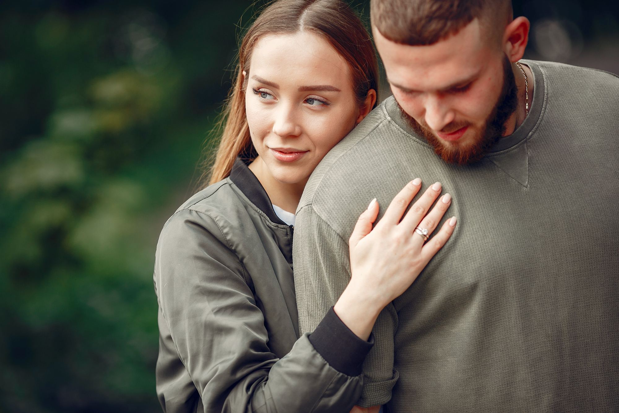 A young woman with long brown hair, wearing a green jacket, embraces a man with short hair and a beard, who is dressed in a gray sweater. They stand outdoors with greenery in the background, both appearing content and close.