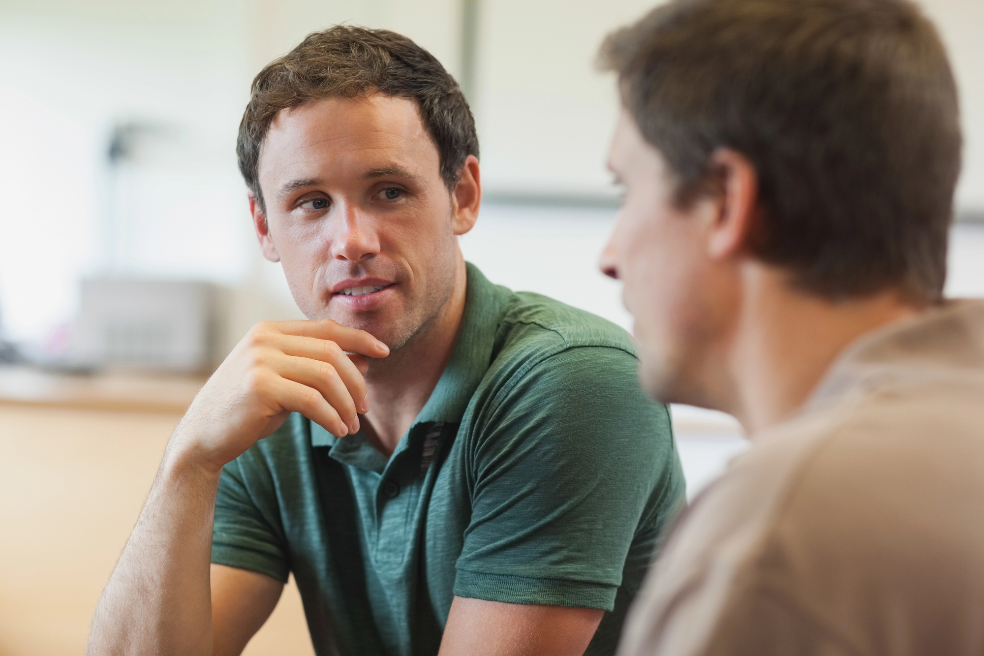 Two men are sitting indoors, engaging in a conversation. The man on the left is wearing a green polo shirt and has a thoughtful expression, resting his hand near his face. The man on the right, slightly blurred, listens attentively. The background is softly lit and out of focus.