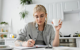 A person with glasses and tattoos, sitting at a table in a modern kitchen, writing in a notebook while holding a white coffee cup. There are kitchen utensils and plants in the background. The person appears focused on their task.