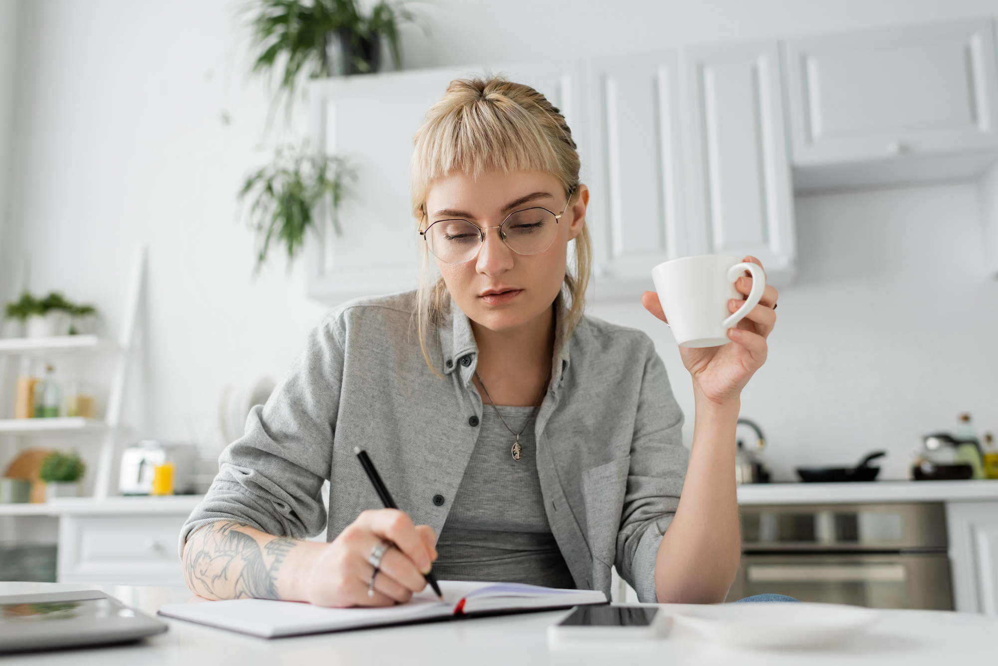 A person with glasses and tattoos, sitting at a table in a modern kitchen, writing in a notebook while holding a white coffee cup. There are kitchen utensils and plants in the background. The person appears focused on their task.