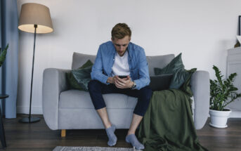 A man is sitting on a gray sofa in a modern living room, intently looking at his smartphone. He is dressed casually in a blue shirt, white t-shirt, and dark pants, with gray socks. A green blanket, a laptop, and some potted plants are nearby. A tall lamp stands in the background.