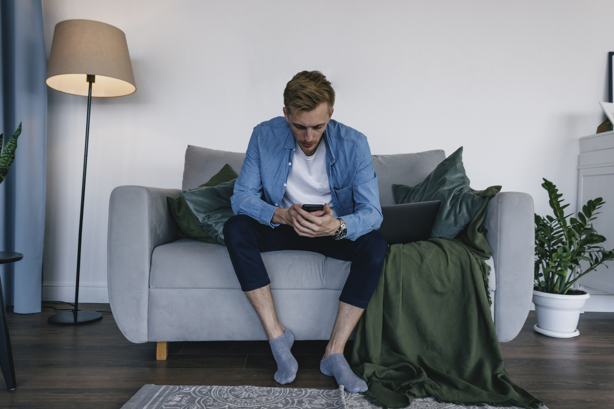 A man is sitting on a gray sofa in a modern living room, intently looking at his smartphone. He is dressed casually in a blue shirt, white t-shirt, and dark pants, with gray socks. A green blanket, a laptop, and some potted plants are nearby. A tall lamp stands in the background.