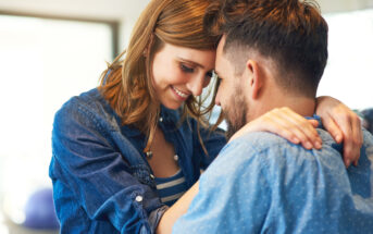 A couple wearing denim shirts are sharing an intimate moment, with their foreheads touching and smiling warmly at each other. The woman has her arms around the man's neck while he gently holds her.