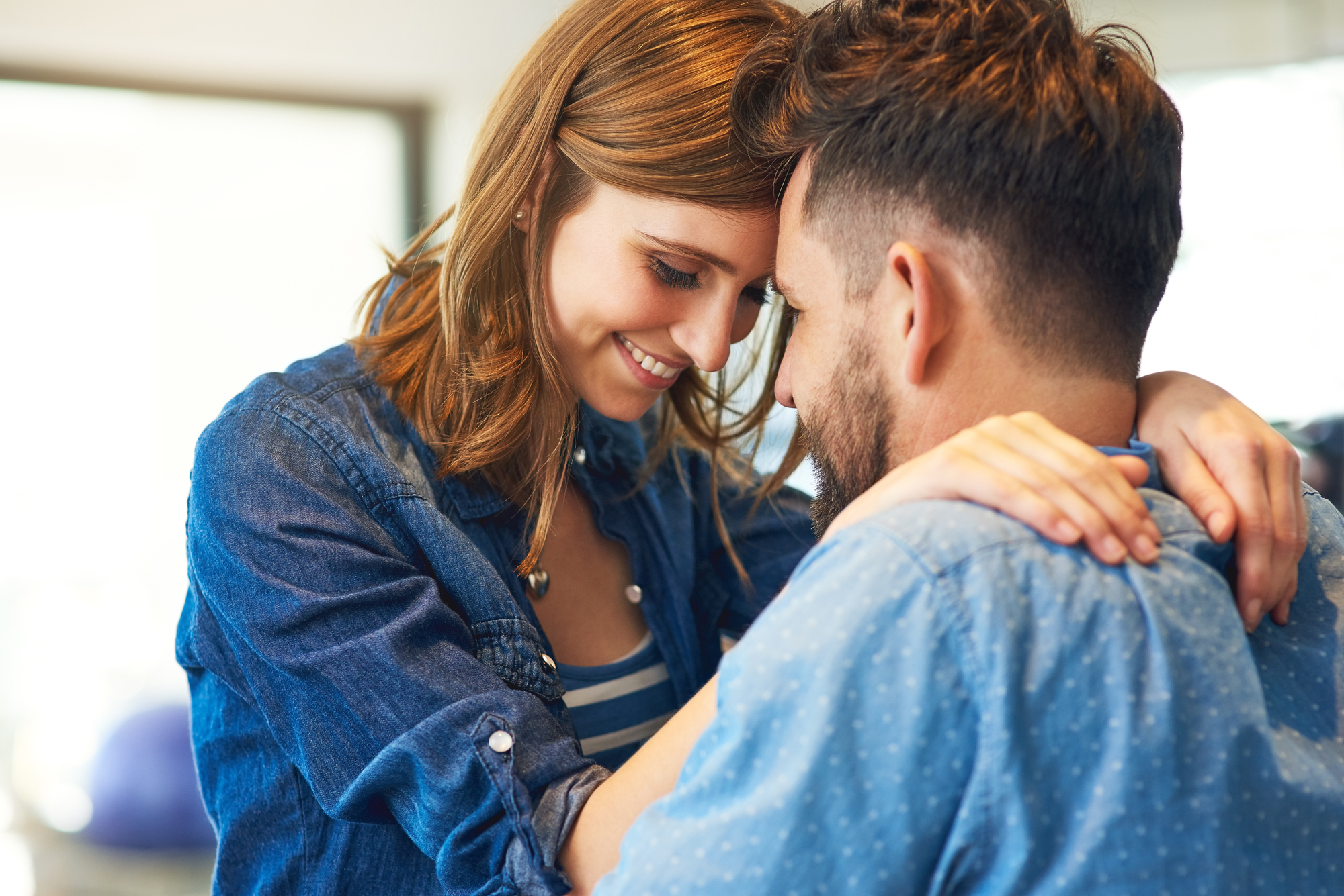 A couple wearing denim shirts are sharing an intimate moment, with their foreheads touching and smiling warmly at each other. The woman has her arms around the man's neck while he gently holds her.