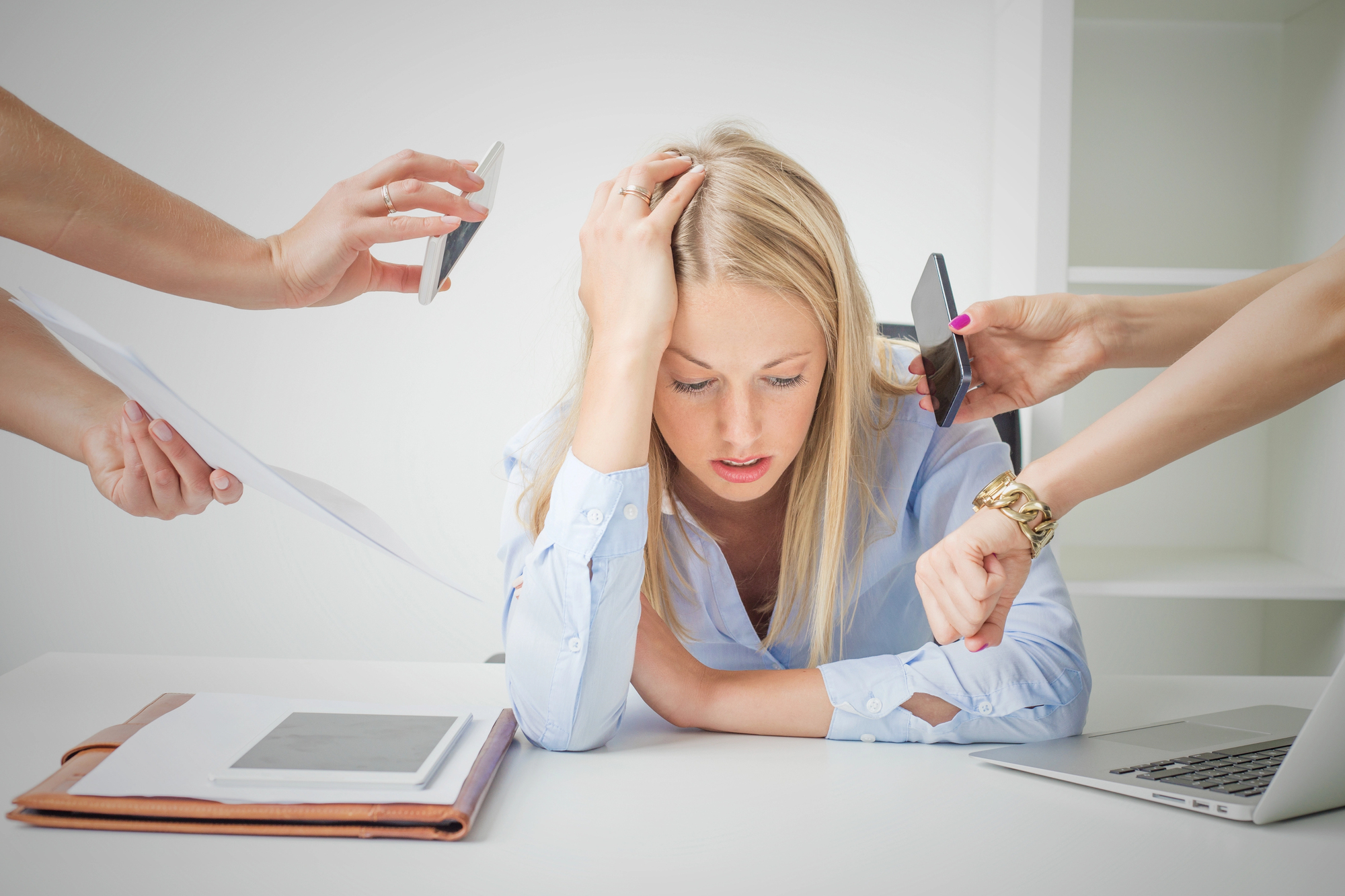 A frustrated woman sits with her head in her hand at a desk, surrounded by several hands holding a phone, a paper, and pointing to a wristwatch, indicating she is overwhelmed with demands. A tablet and laptop are on the desk.