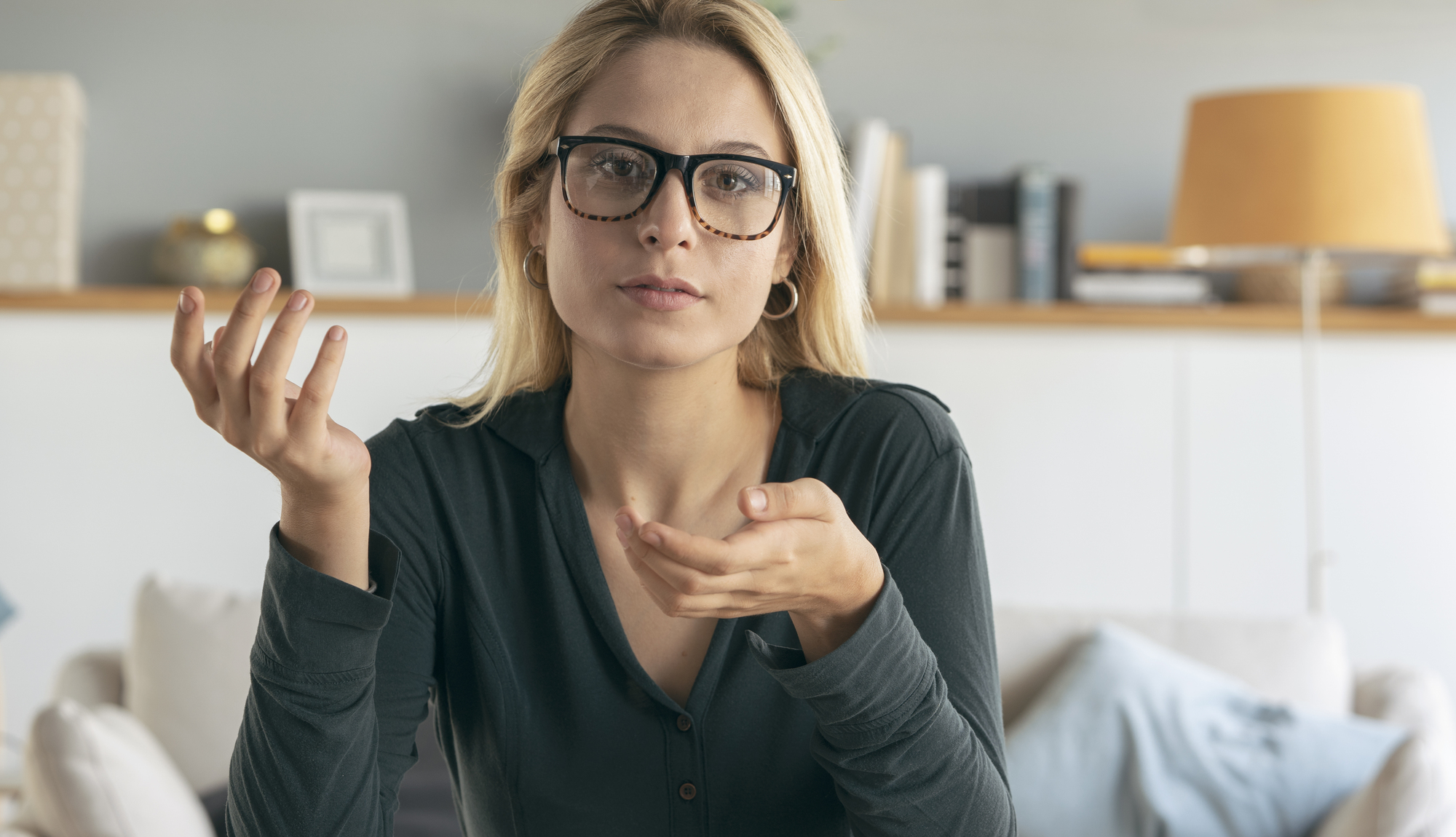 A woman with long blonde hair and glasses is sitting on a couch in a living room, looking at the camera with a questioning expression. She is wearing a dark long-sleeved shirt, and books and a lamp can be seen in the background.