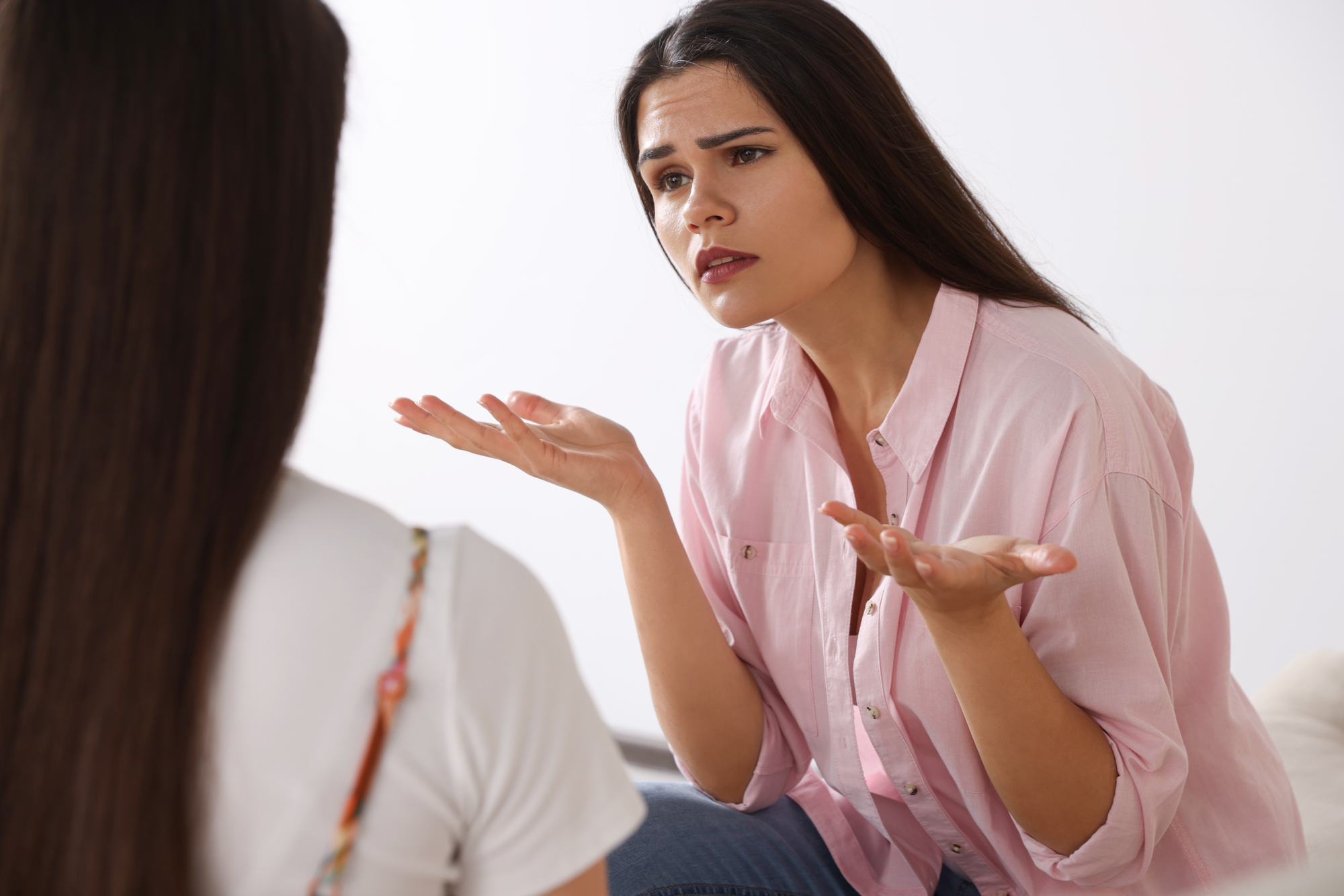 A woman with long dark hair, wearing a pink shirt, leans forward with a perplexed expression, hands outstretched and palms up. She appears to be speaking to another person whose back is to the camera, only partially visible on the left side of the image.