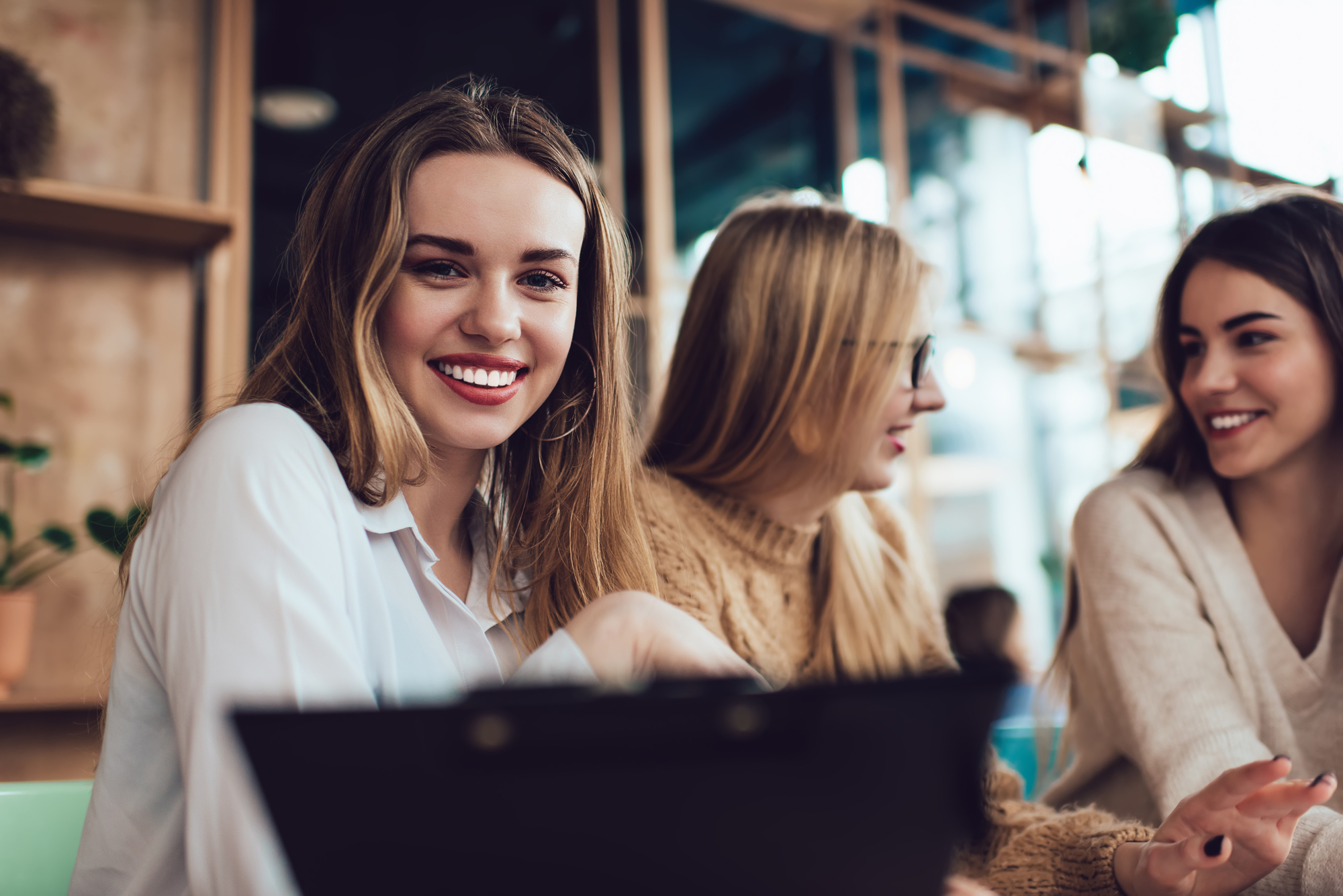 Three young women are seated in a cafe, engaged in conversation. One woman in the foreground, wearing a white blouse, is smiling at the camera. The other two women, wearing sweaters, are slightly blurred in the background, focusing on their conversation.