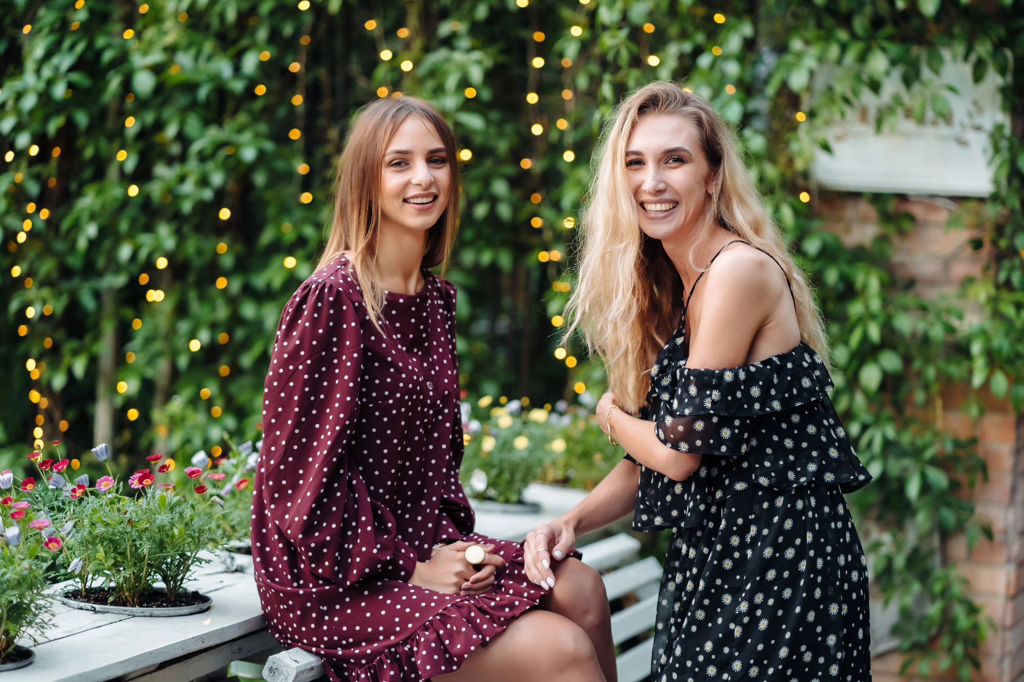 Two women are enjoying an outdoor setting with lush green foliage and string lights in the background. Both are wearing patterned dresses and smiling at the camera. One woman is seated on a bench while the other stands next to her. Decorative flowers surround them.