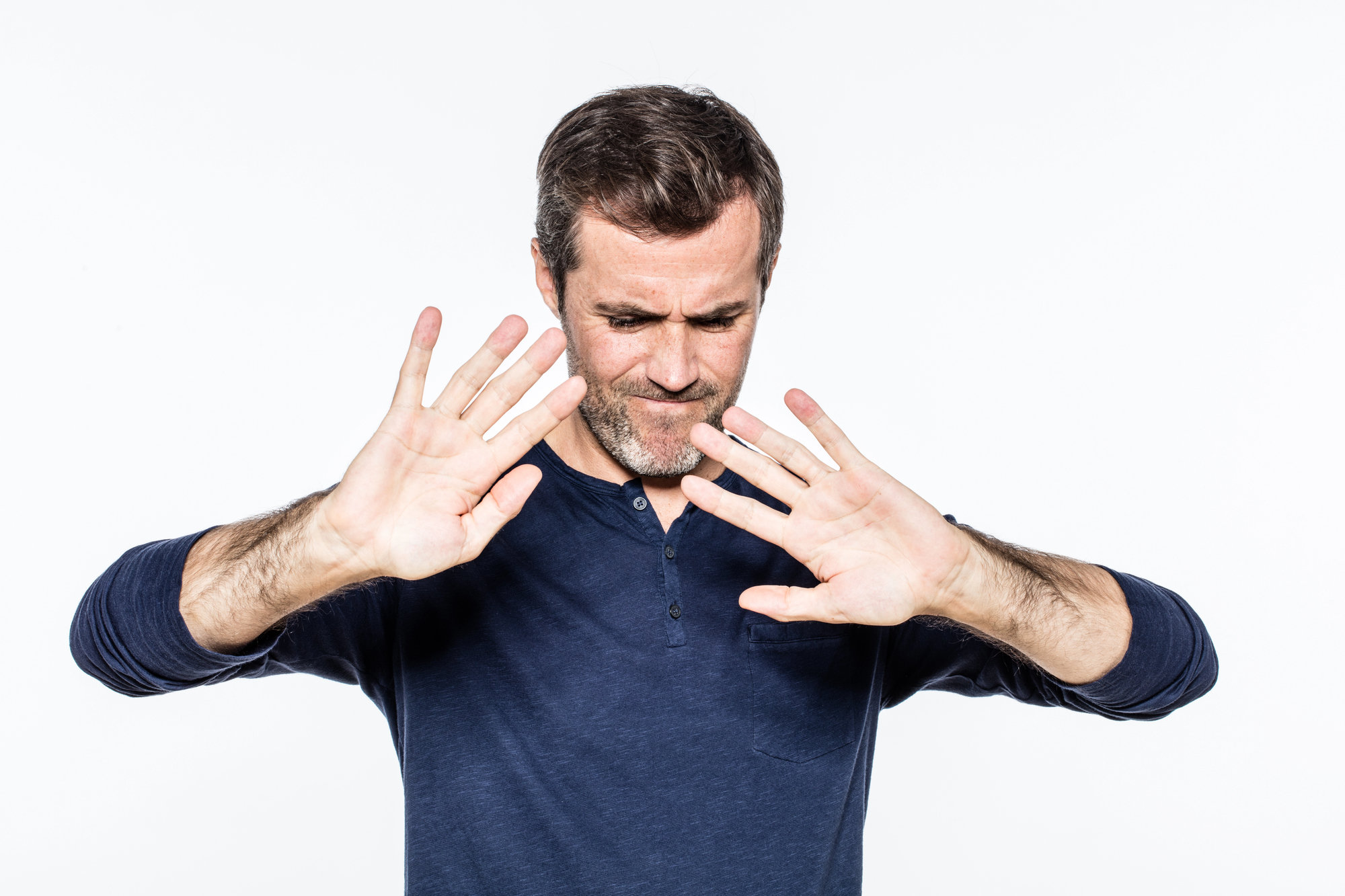 A middle-aged man with short hair and a beard, wearing a navy blue shirt, holds his hands up in front of him as if to protect himself or gesture "stop." He is looking downward with a concerned expression. The background is plain white.