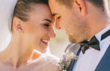 A close-up of a bride and groom touching their foreheads together and smiling lovingly. The bride wears a white veil and lace dress, and the groom is in a gray suit with a black bow tie and a small floral boutonniere.