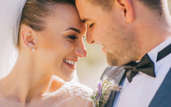 A close-up of a bride and groom touching their foreheads together and smiling lovingly. The bride wears a white veil and lace dress, and the groom is in a gray suit with a black bow tie and a small floral boutonniere.