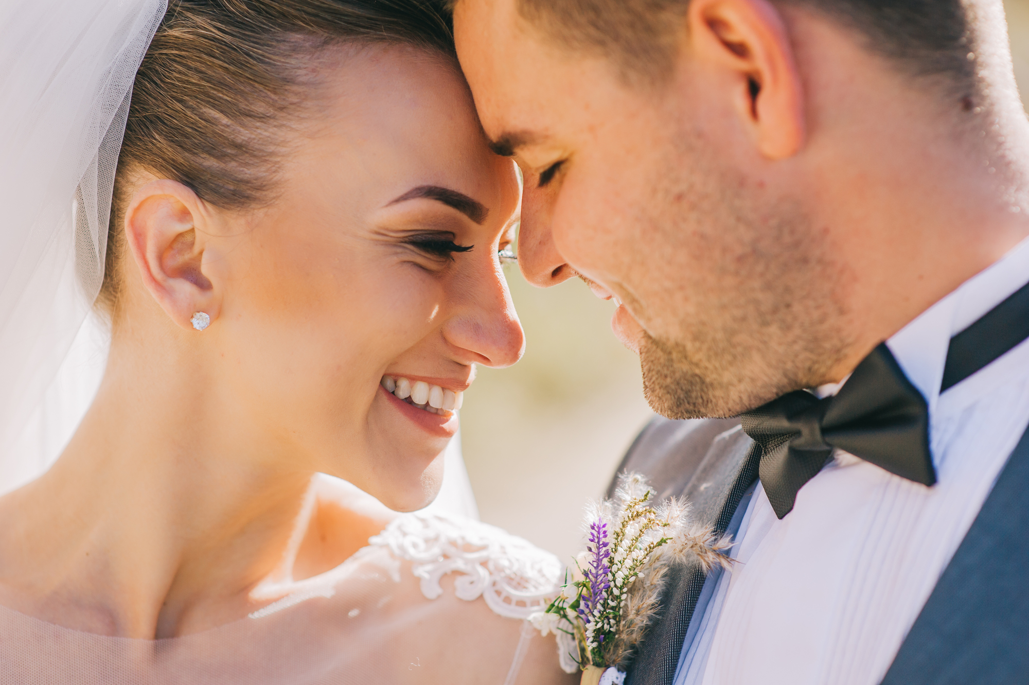 A close-up of a bride and groom touching their foreheads together and smiling lovingly. The bride wears a white veil and lace dress, and the groom is in a gray suit with a black bow tie and a small floral boutonniere.