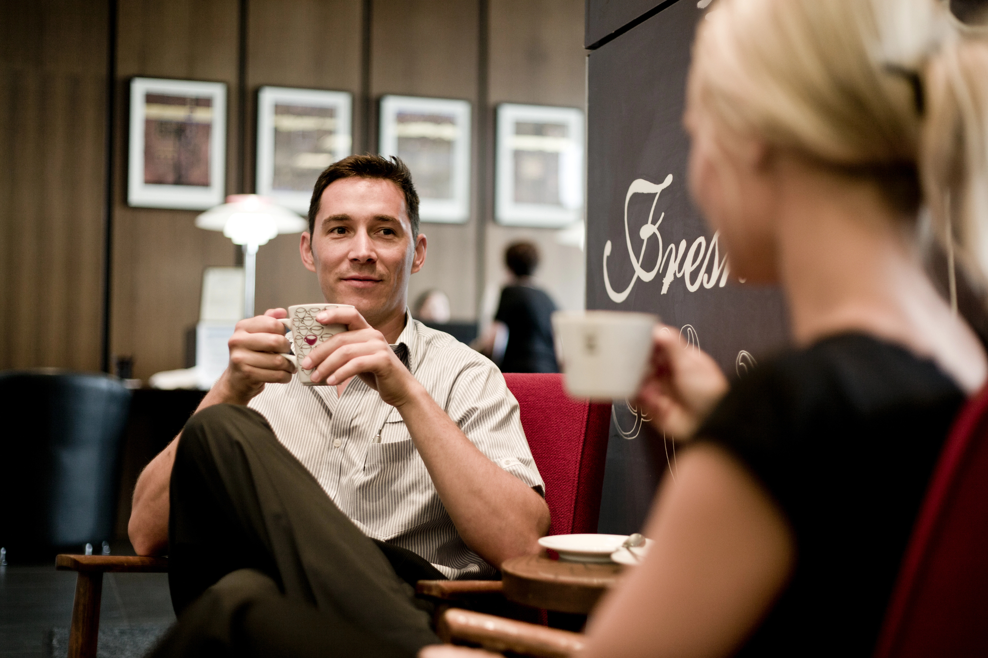 A man and a woman are sitting in a cozy cafe, each holding a cup of coffee. The man, dressed in a light shirt and dark pants, is smiling and making eye contact with the woman, who is slightly out of focus in the foreground. The cafe has a warm, inviting atmosphere.