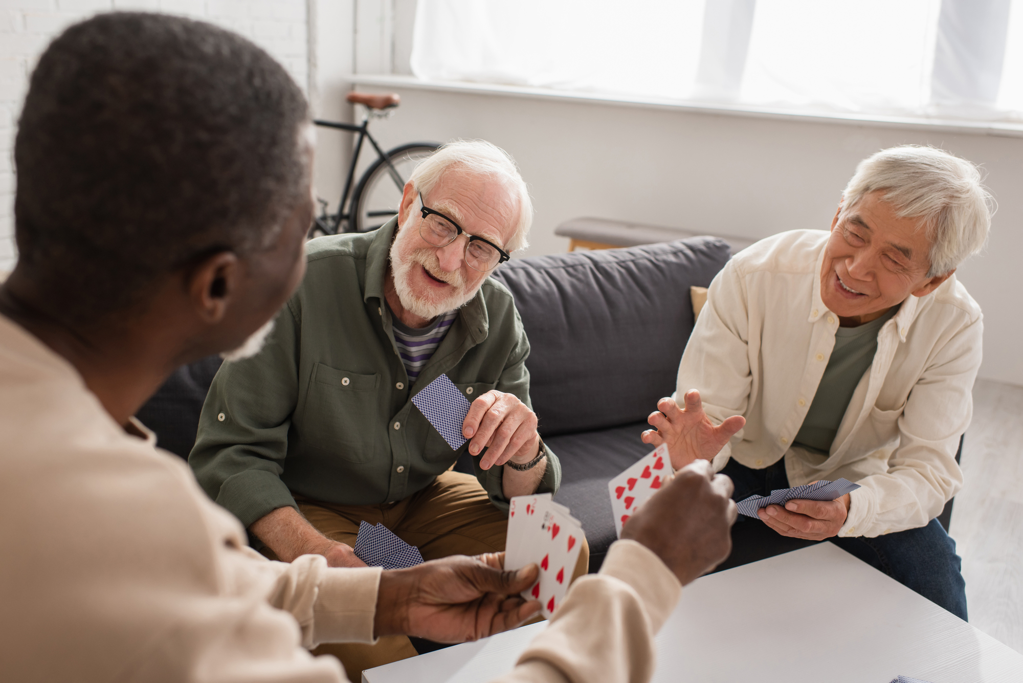 Three elderly friends sit together at a table in a bright room, playing a card game. They are smiling and appear to be enjoying each other's company. A bicycle is visible in the background near a couch and window.