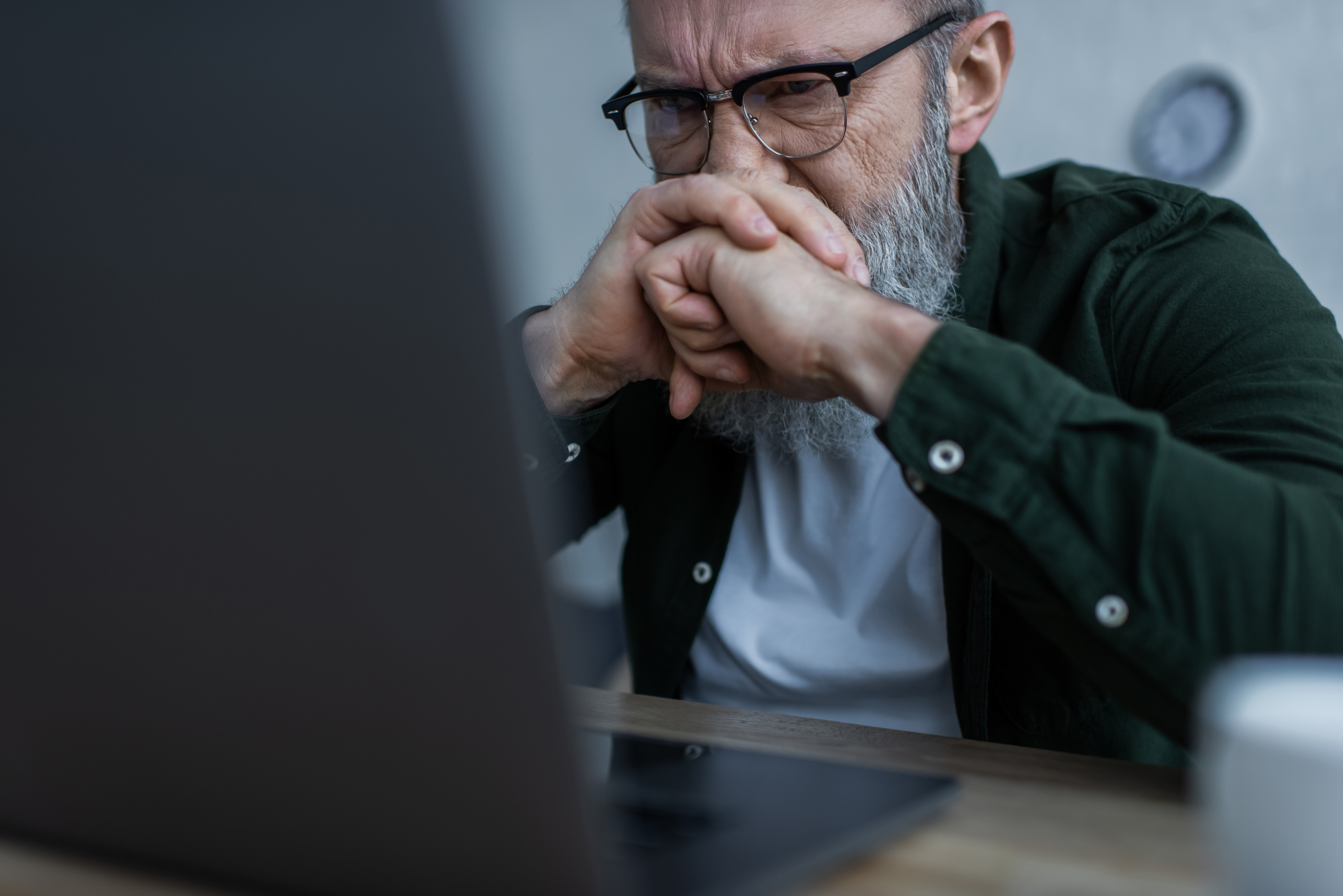 A man with a beard, glasses, and a green shirt sits at a desk, looking distressed while staring at a laptop screen. He has his hands clasped in front of his mouth, with furrowed brows and closed eyes, suggesting concentration or worry.