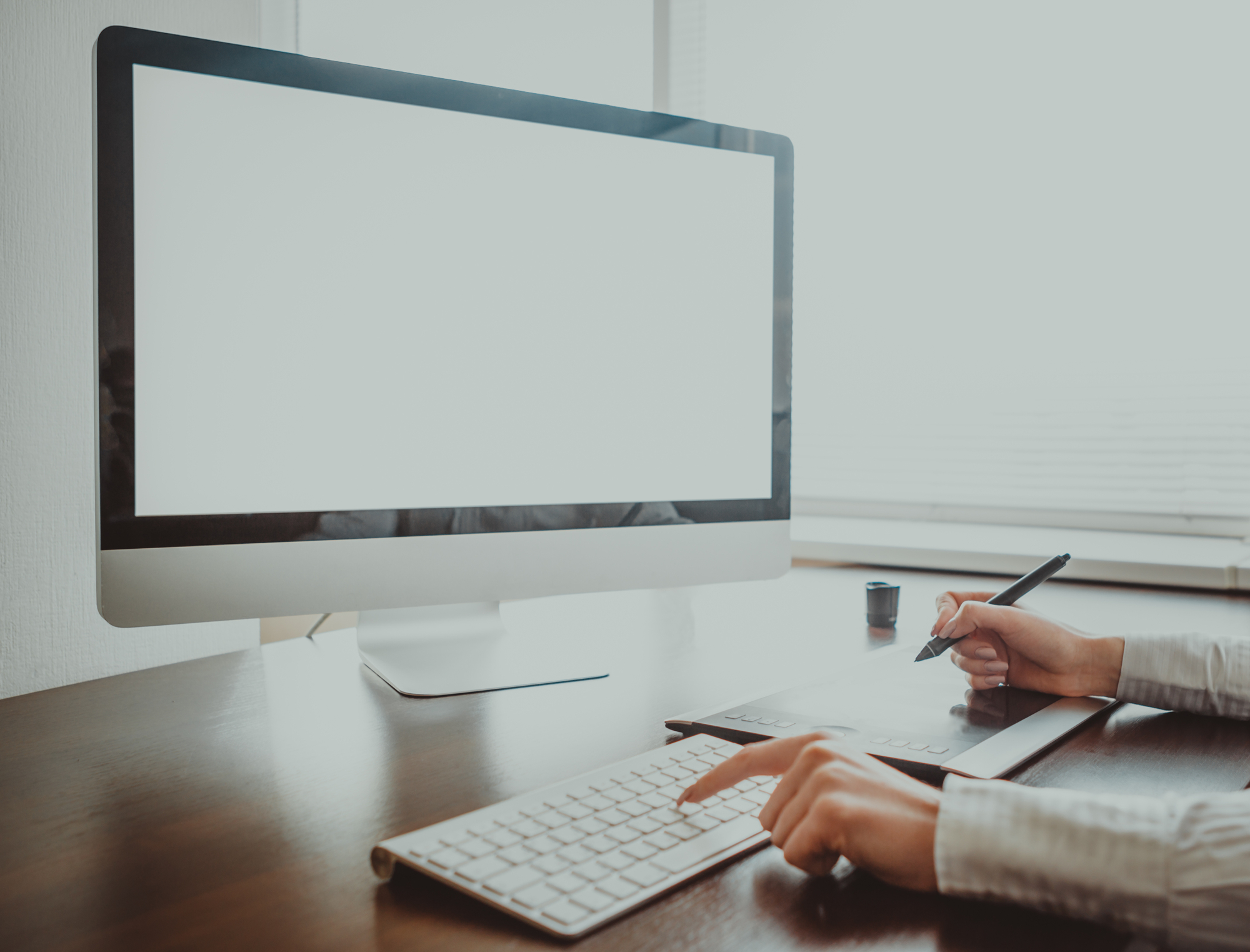 A person is working at a sleek wooden desk with a large computer monitor displaying a blank white screen. One hand is using a stylus on a drawing tablet, while the other hand is typing on a white keyboard. The workspace is well-lit with natural light.