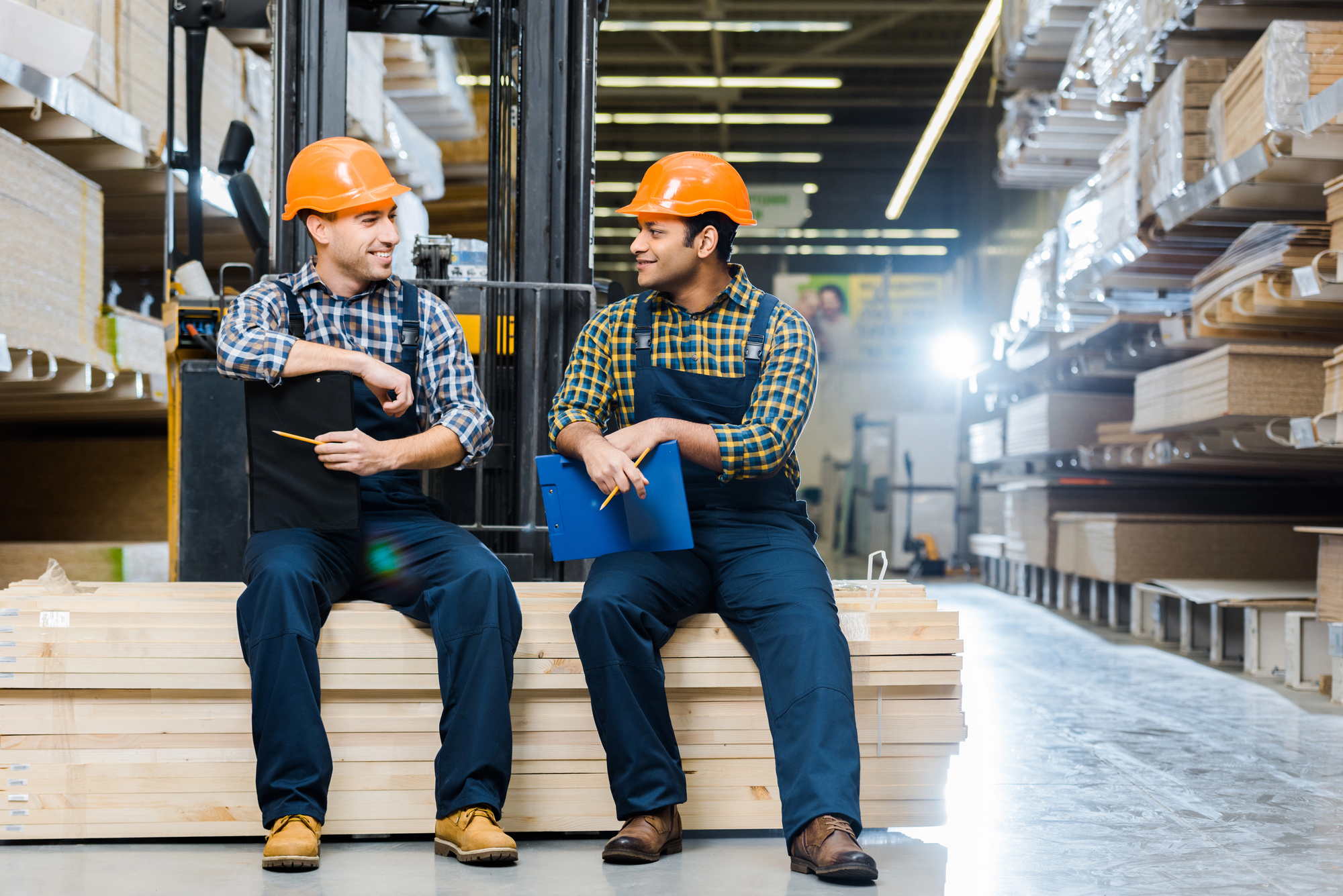 Two workers in safety helmets and plaid shirts sit on a stack of wooden planks, taking a break in a warehouse. They hold clipboards and smile at each other. Shelves stocked with materials line the background, and a forklift is partially visible behind them.