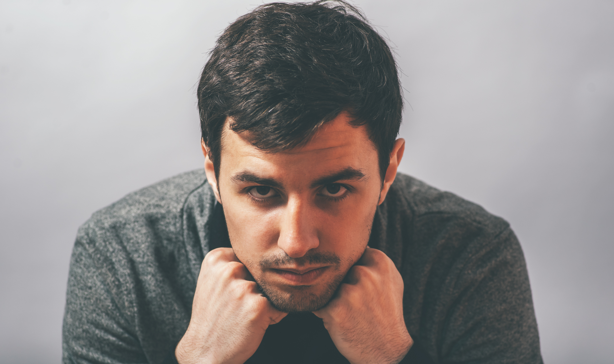 A man with short, dark hair and stubble stares intensely at the camera, resting his chin on his folded hands. He is wearing a dark gray sweater and is set against a plain gray background.