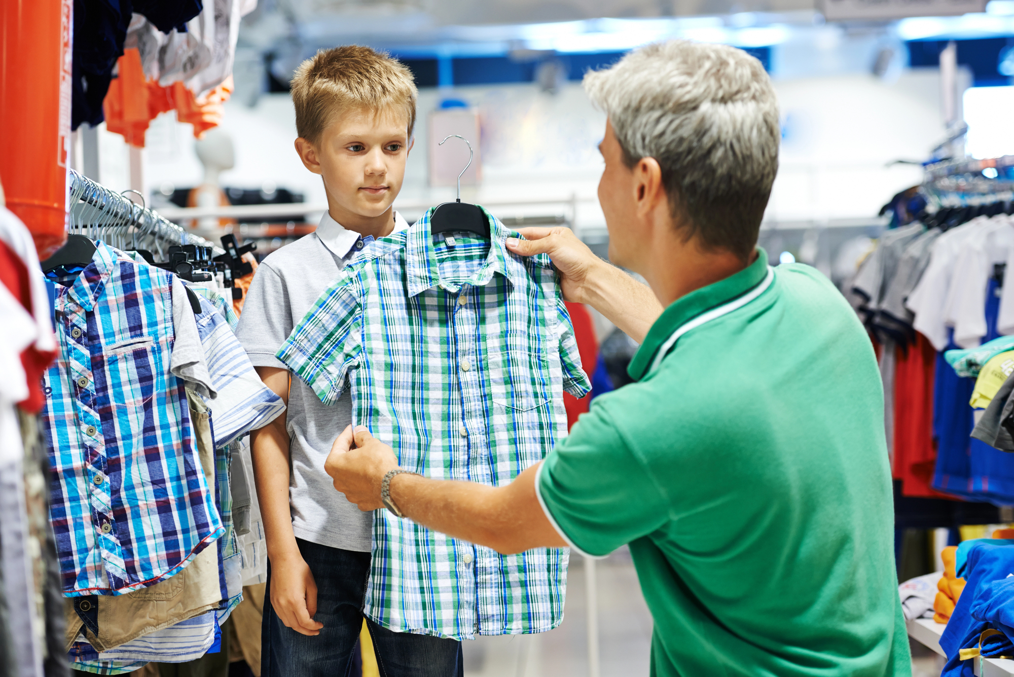 An adult is shopping for clothes with a young boy in a store. The adult is holding up a green and blue plaid shirt on a hanger, showing it to the boy. They are surrounded by various shirts and clothing items.