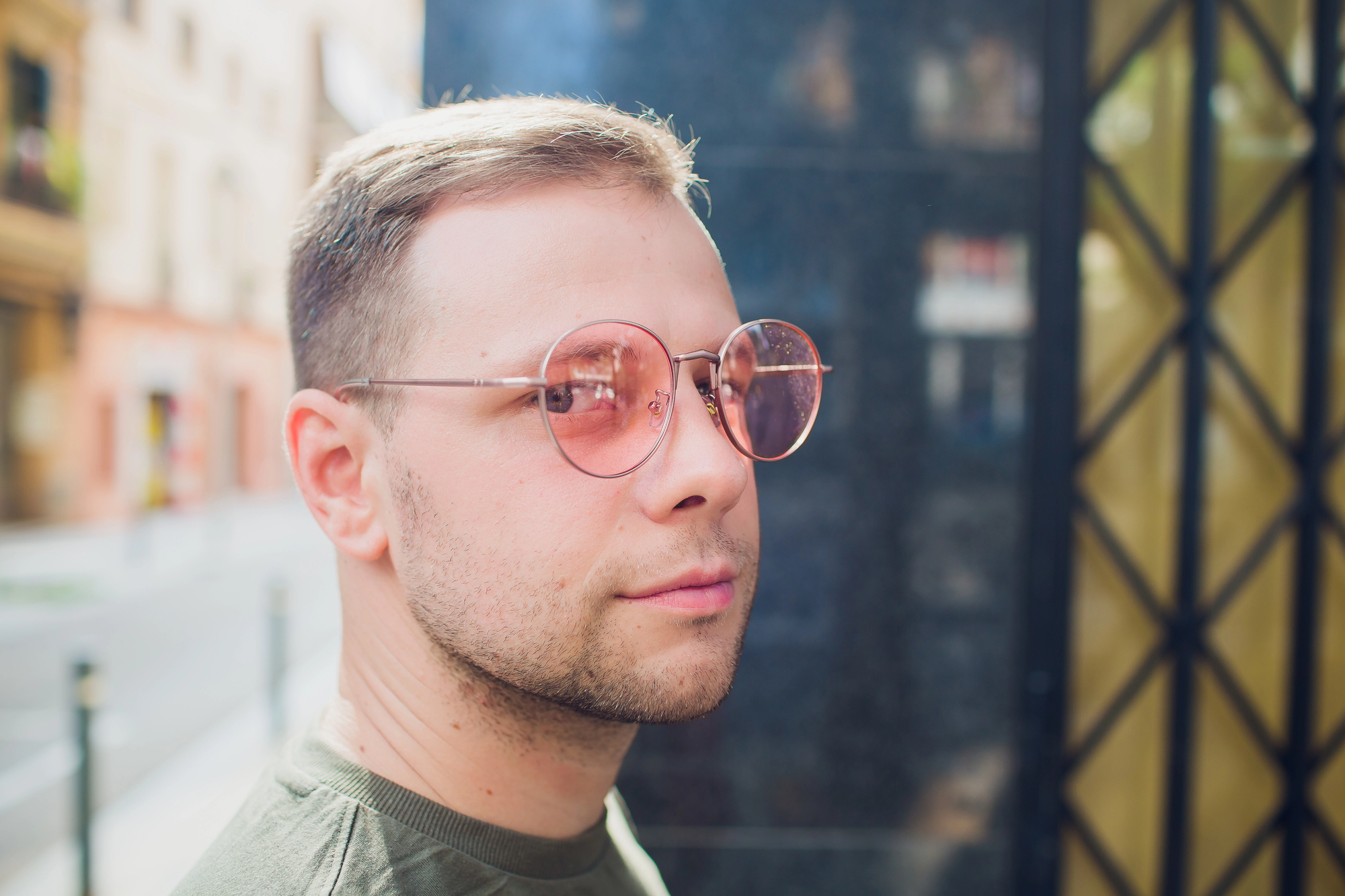 A man with short hair and a trimmed beard, wearing round, light-pink tinted glasses, is looking sideways at the camera. He is outdoors, with urban buildings and a blurred street scene in the background.