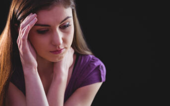 A woman with long brown hair, wearing a purple shirt, appears deep in thought or worried. She rests one hand on her forehead and the other near her neck. The background is dark, emphasizing her contemplative expression.