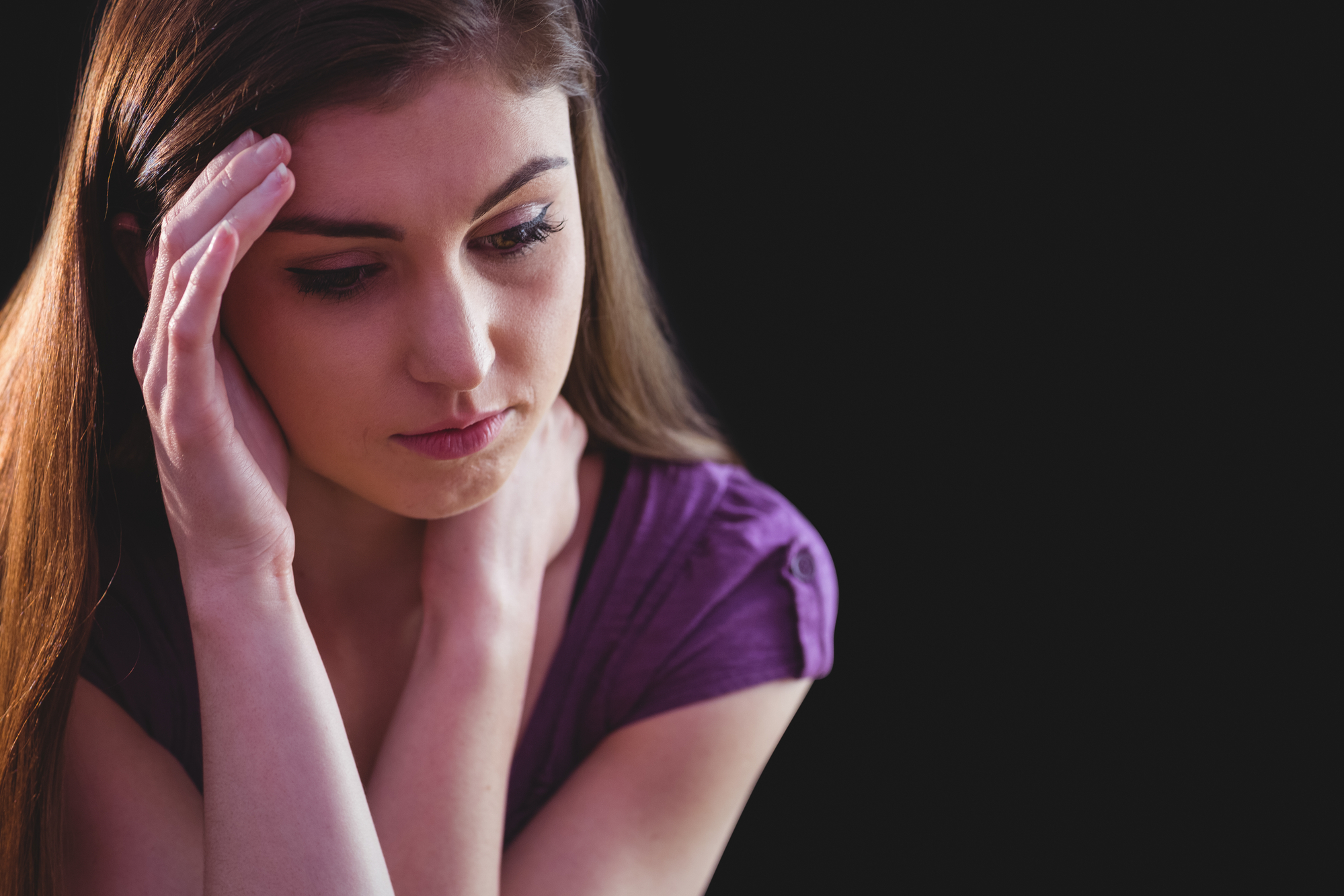 A woman with long brown hair, wearing a purple shirt, appears deep in thought or worried. She rests one hand on her forehead and the other near her neck. The background is dark, emphasizing her contemplative expression.