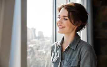 A woman with short brown hair smiles while looking out a window. She is wearing a gray button-up shirt, and sunlight illuminates her face. Blurred cityscape is visible outside the window.