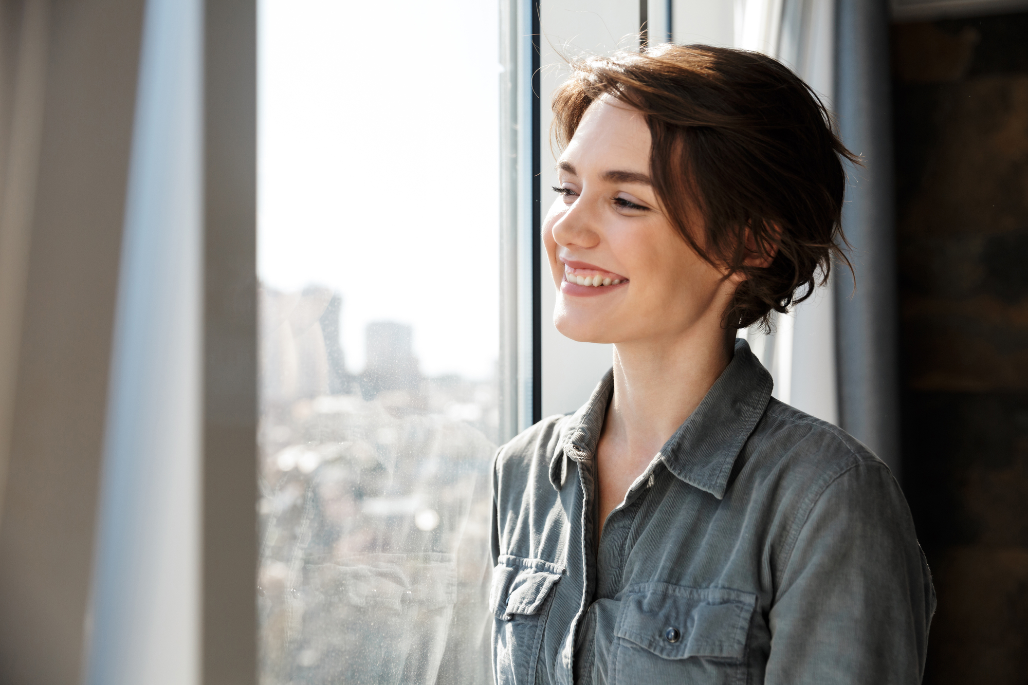 A woman with short brown hair smiles while looking out a window. She is wearing a gray button-up shirt, and sunlight illuminates her face. Blurred cityscape is visible outside the window.