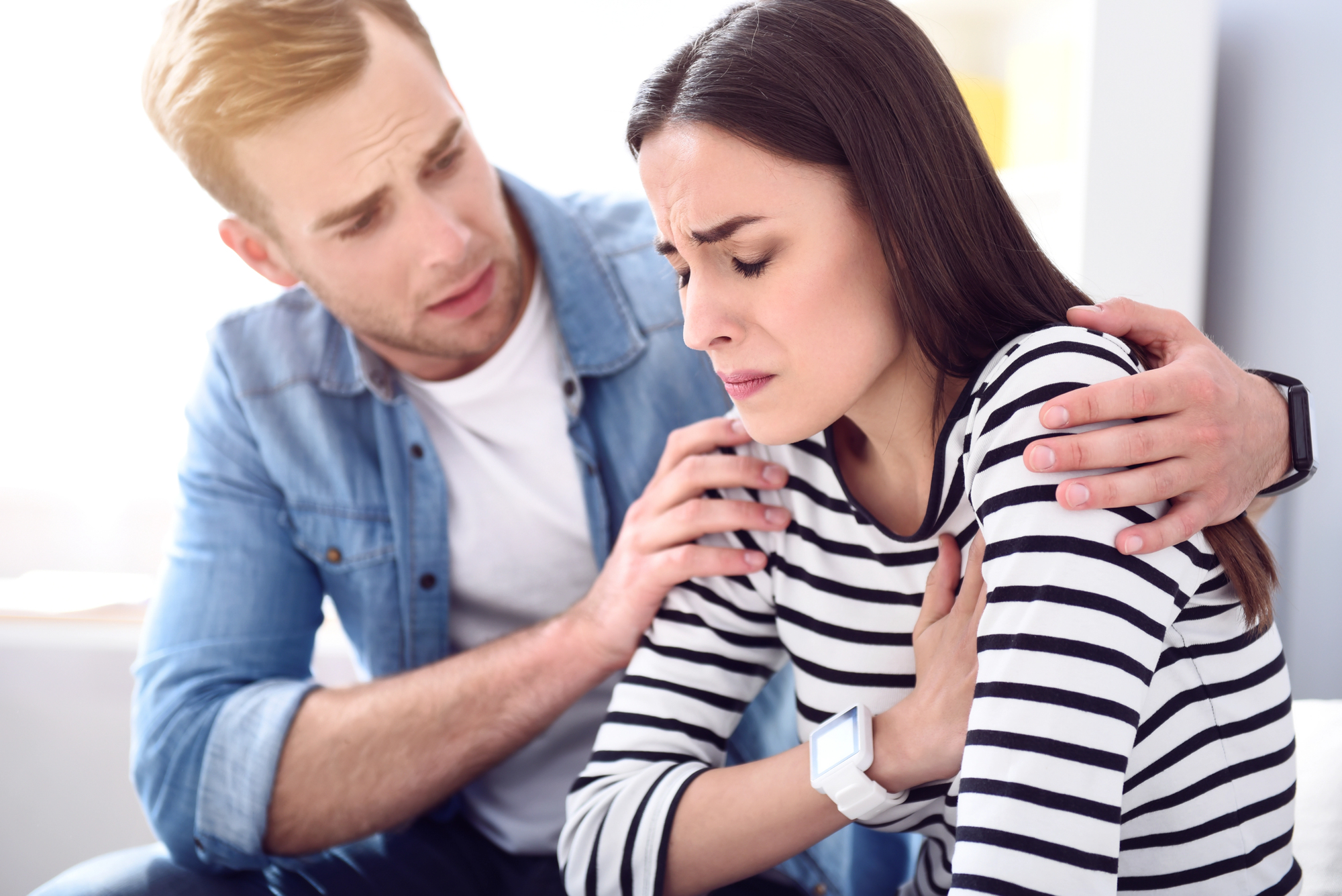 A concerned man in a blue denim shirt places his hands on the shoulders of a woman in a striped shirt, who appears to be in distress. She has her hand on her chest and looks away, with a pained expression on her face.