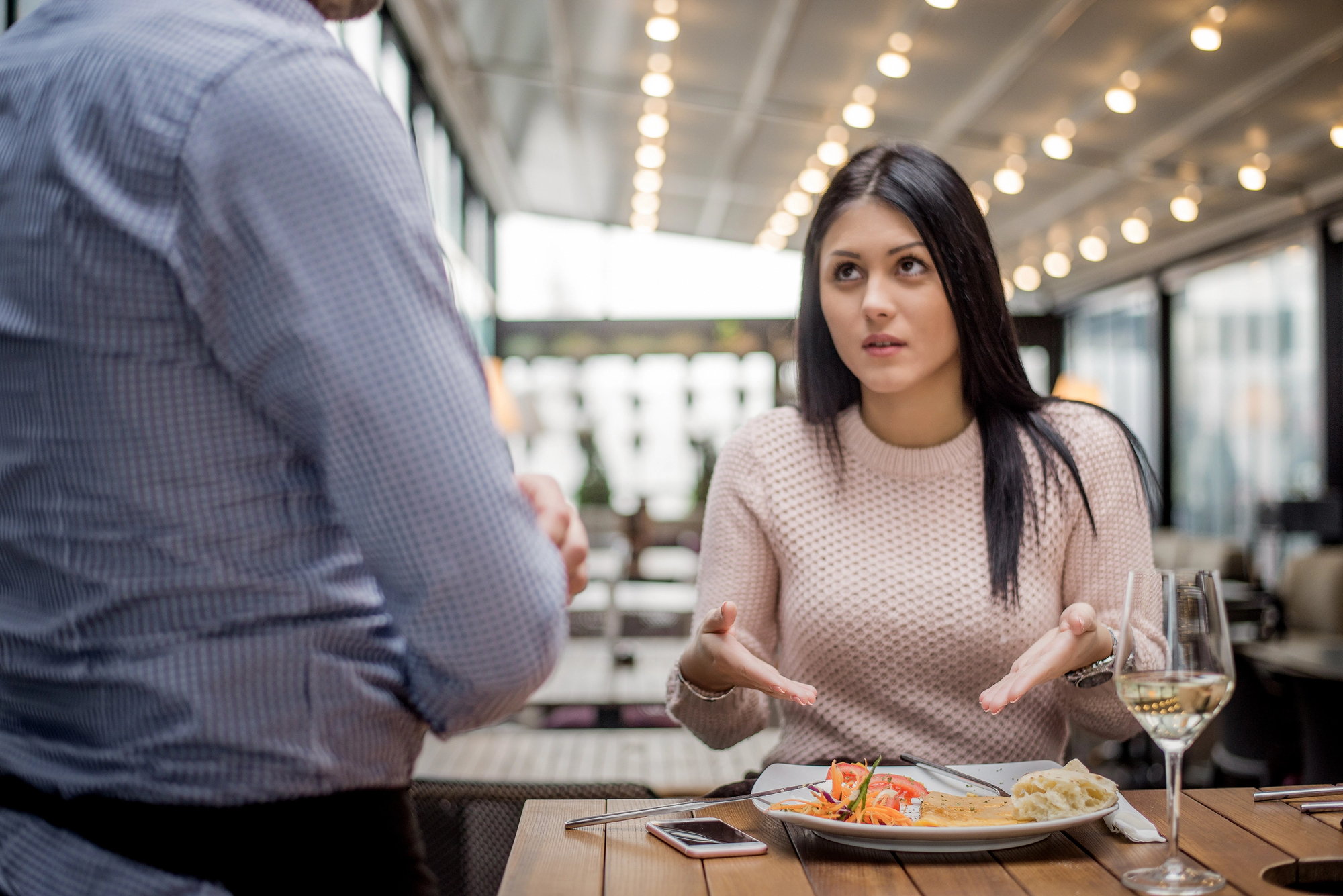 A woman with long dark hair in a pink sweater is sitting at a restaurant table, gesturing with her hands while speaking to a person standing nearby. The table has a plate of food, a glass of white wine, and a smartphone on it. String lights are visible in the background.