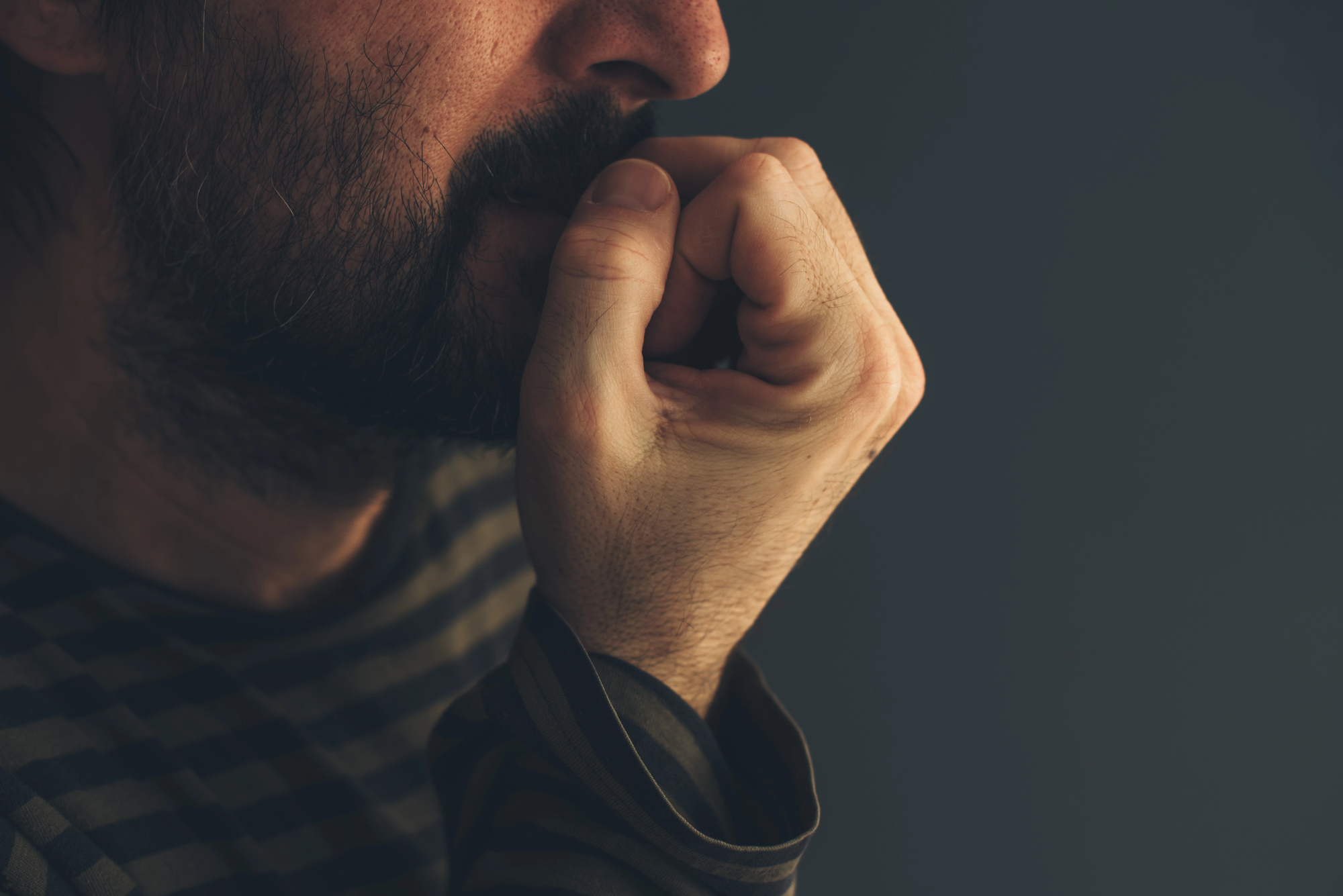 Close-up of a person with a beard, appearing to be in deep thought or concern. They rest their chin on their hand, with fingers partially covering their mouth. The background is dark and blurred, drawing focus to the person's contemplative expression.