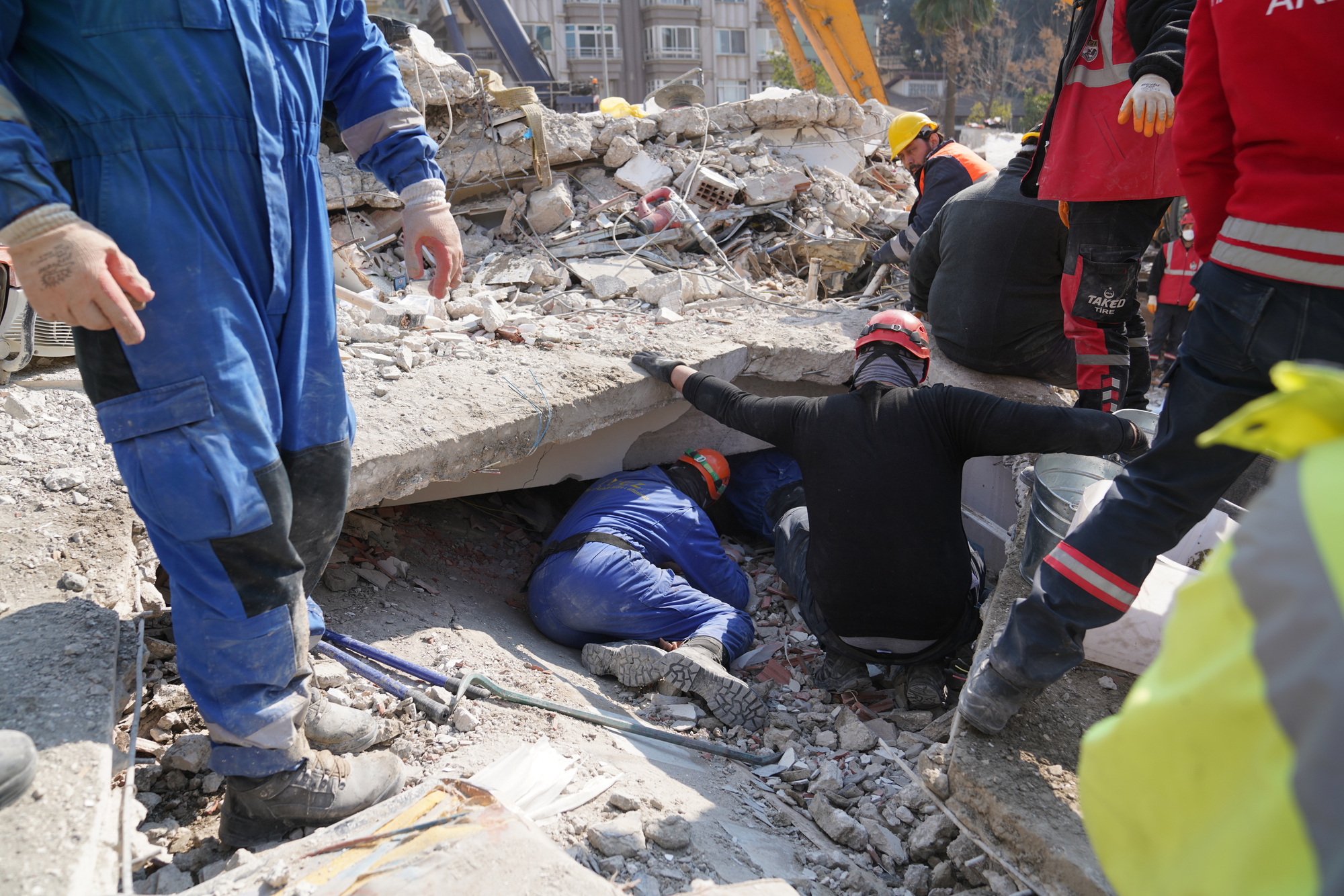 Rescue workers in protective gear search for survivors under collapsed concrete slabs amidst rubble, following a structural collapse in an urban area. Construction machinery and support personnel are visible in the background, coordinating the effort.