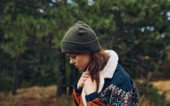 A person with long brown hair and a green beanie looks down while standing in an outdoor setting with greenery and trees in the background. They are wearing a colorful patterned sweater with a sherpa lining.