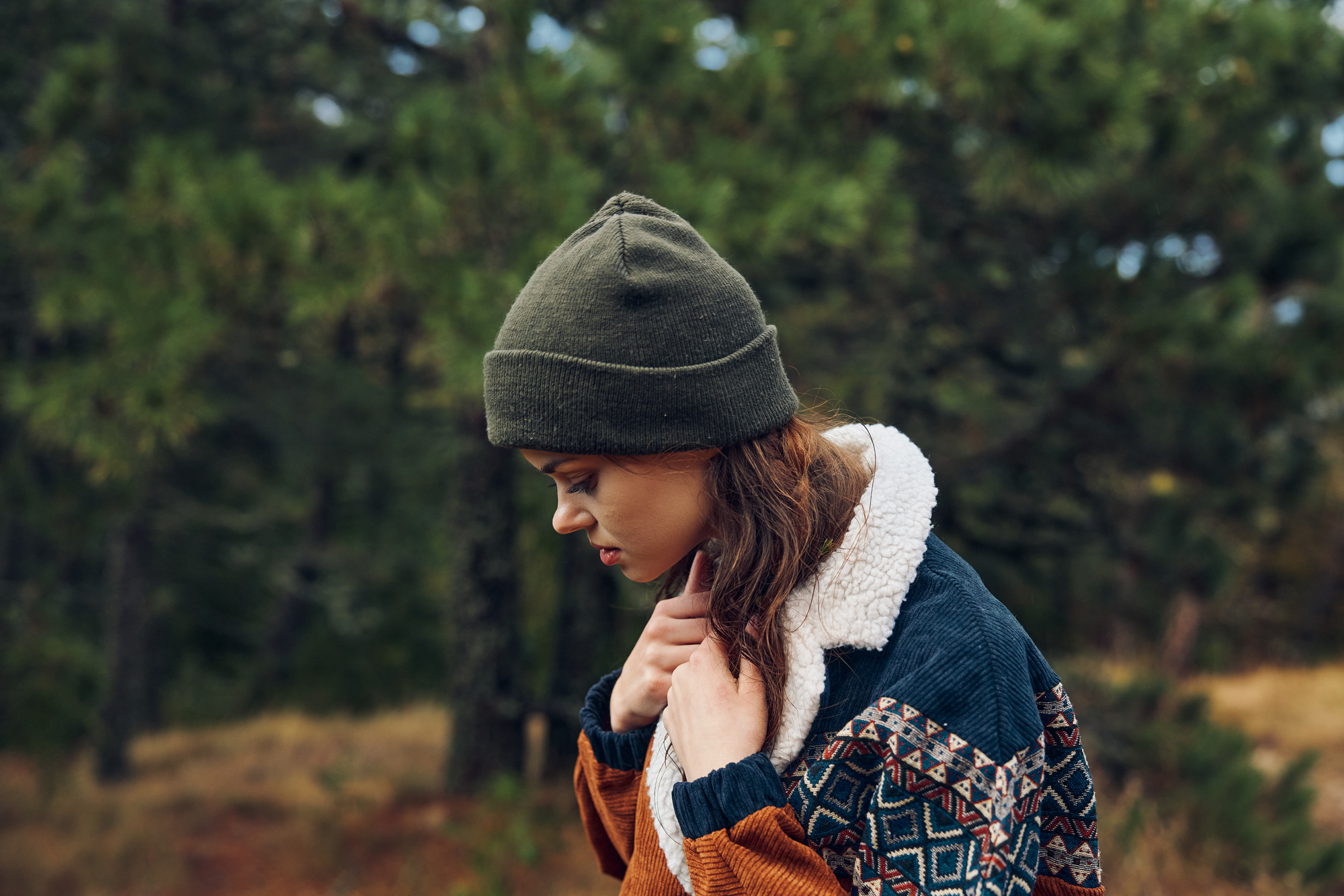 A person with long brown hair and a green beanie looks down while standing in an outdoor setting with greenery and trees in the background. They are wearing a colorful patterned sweater with a sherpa lining.