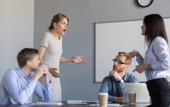 A group of four colleagues are in a heated discussion at a conference table. One person is standing with arms outstretched, appearing frustrated. The others are seated, one with crossed arms and another holding a document. There are coffee cups and a laptop on the table.