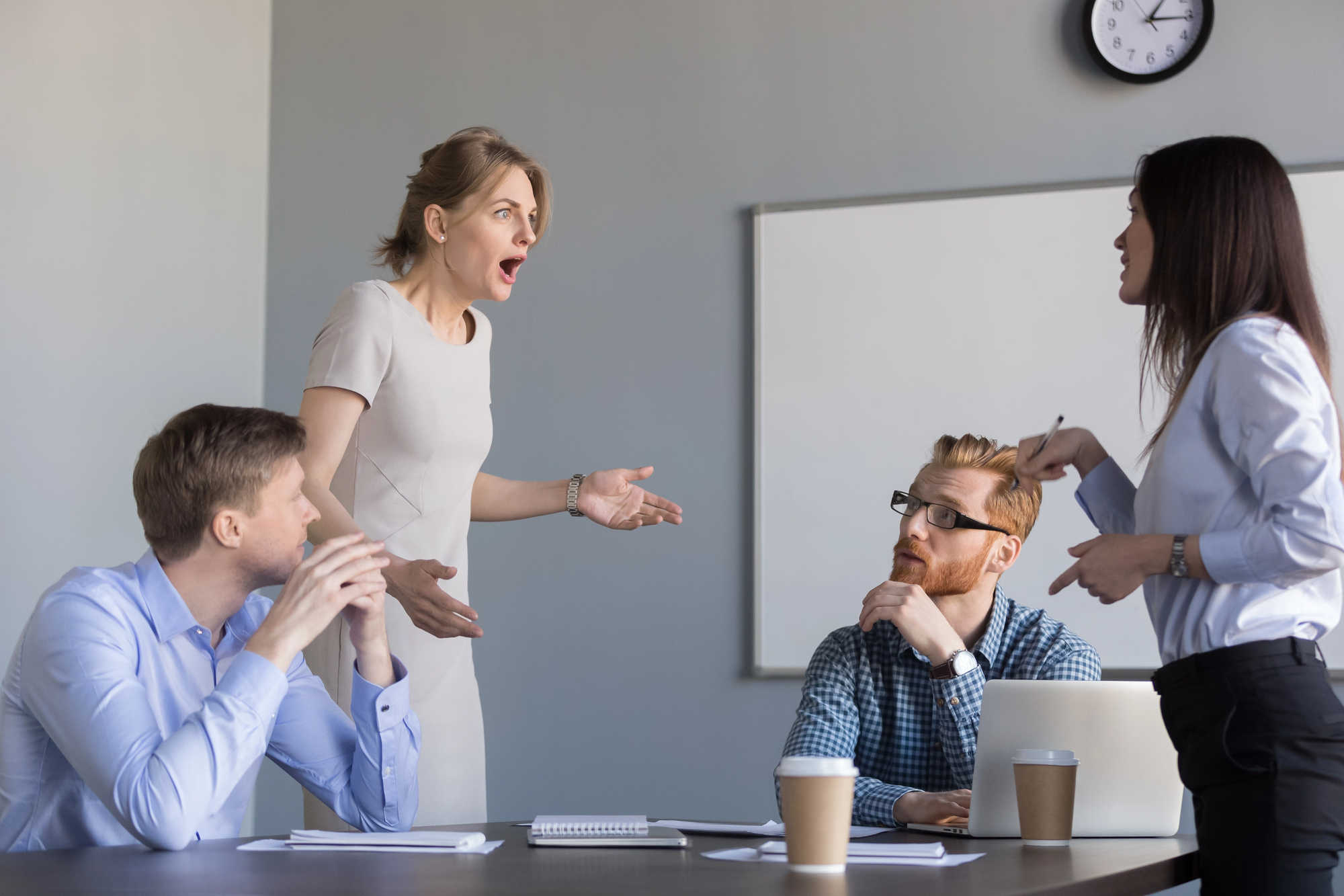 A group of four colleagues are in a heated discussion at a conference table. One person is standing with arms outstretched, appearing frustrated. The others are seated, one with crossed arms and another holding a document. There are coffee cups and a laptop on the table.