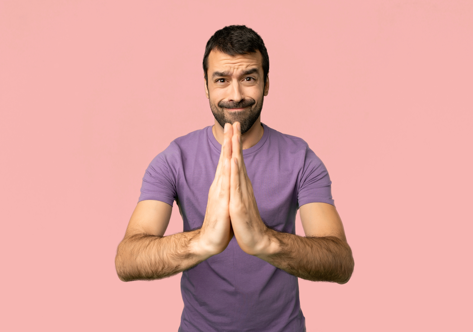 A man with a beard and mustache wearing a purple t-shirt stands against a pink background. He is holding his hands together in a prayer-like gesture and smiling with one eyebrow slightly raised, giving a playful or slightly confused expression.