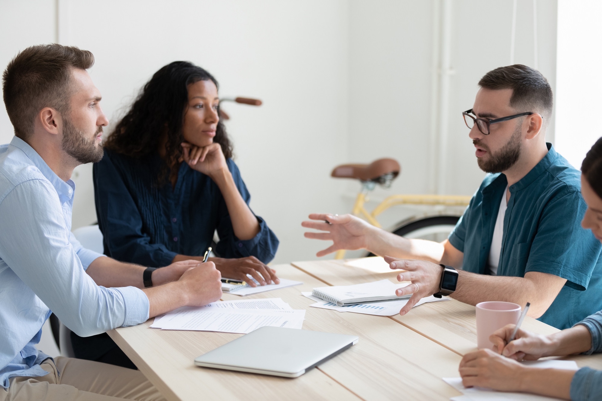 A group of four people are sitting around a table in a meeting room, engaged in a discussion. One person gestures while speaking, and the others listen attentively. Papers, a laptop, and a pink coffee mug are on the table. A bicycle is visible in the background.