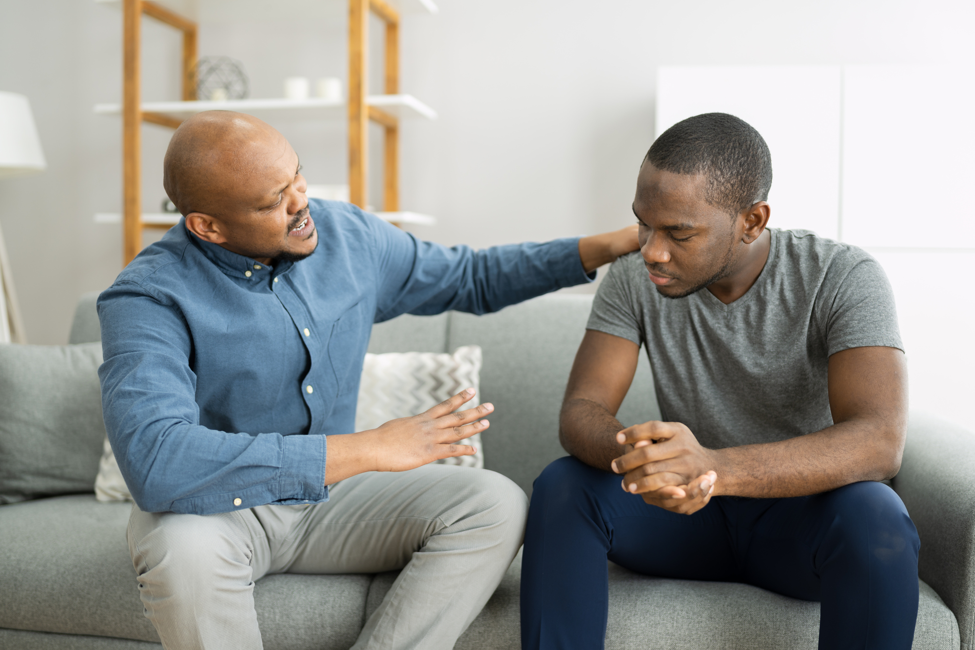 Two men sitting on a couch engaged in a serious conversation. One man, dressed in a blue shirt, is gesturing with his right hand while resting his left hand on the other man's shoulder. The second man, wearing a gray t-shirt, is looking down with his hands clasped.