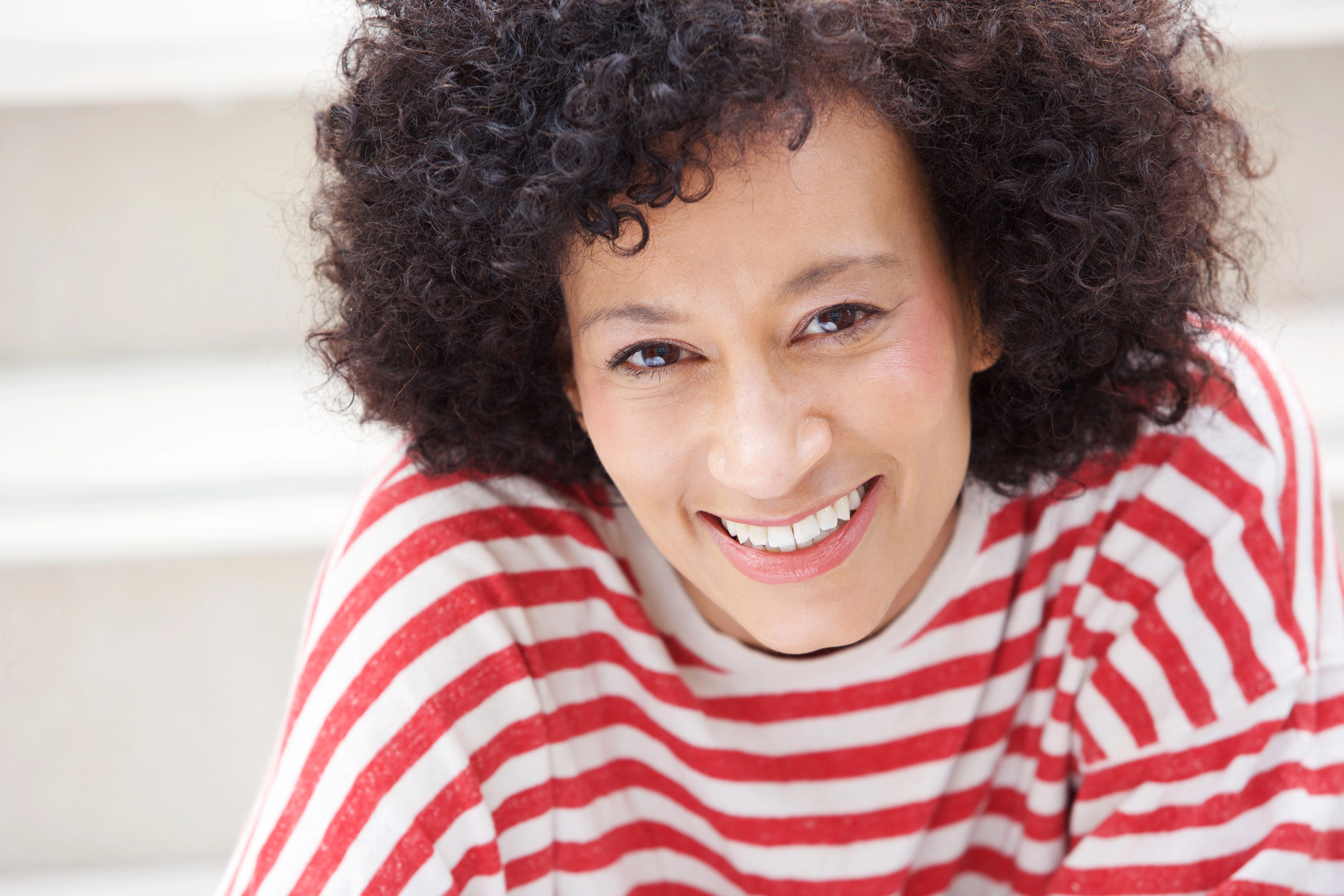 A person with curly hair smiles warmly at the camera. They are wearing a red and white striped shirt and are outdoors, with blurry white steps in the background.