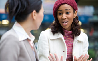 Two women are engaged in conversation outdoors. The woman on the left, with dark hair tied back, is facing another woman who has a curly hair, dressed in a white coat and a red knitted hat. The background is slightly blurred, hinting at an urban setting.