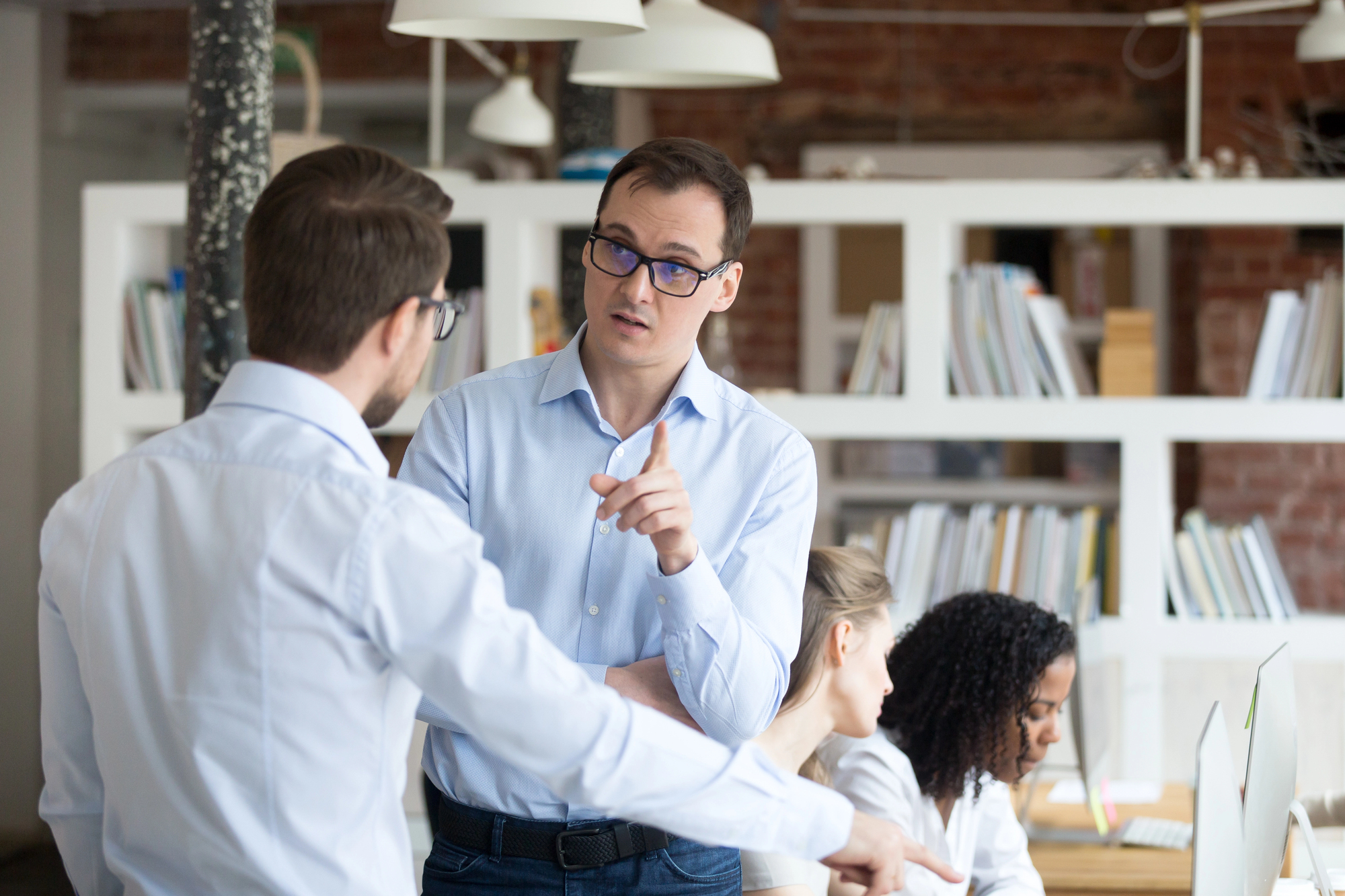 A man in a blue shirt and glasses is pointing and talking to another man with glasses in an office setting. In the background, three people are working at computers near a bookshelf filled with various items. The setting has a modern, open-plan design.