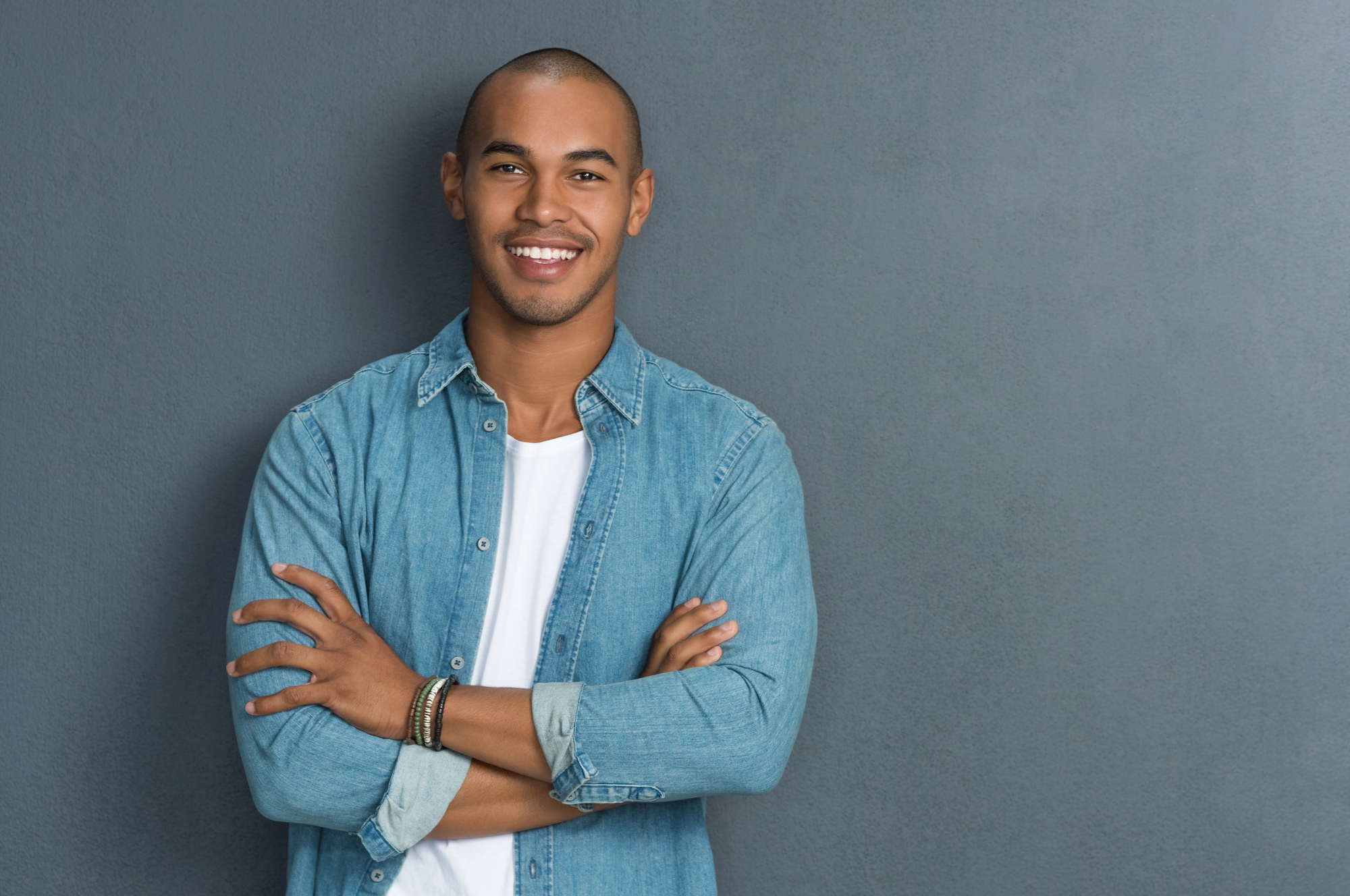 A smiling person with a shaved head is standing against a gray wall with their arms crossed. They are wearing a blue denim shirt over a white t-shirt and bracelets on their left wrist.